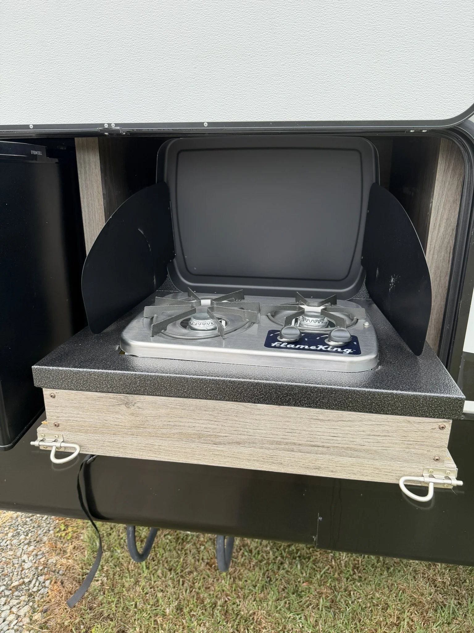A portable stove with two burners in a compartment of a camper or RV, with a black cover and a wooden exterior, on a patch of grass and gravel.