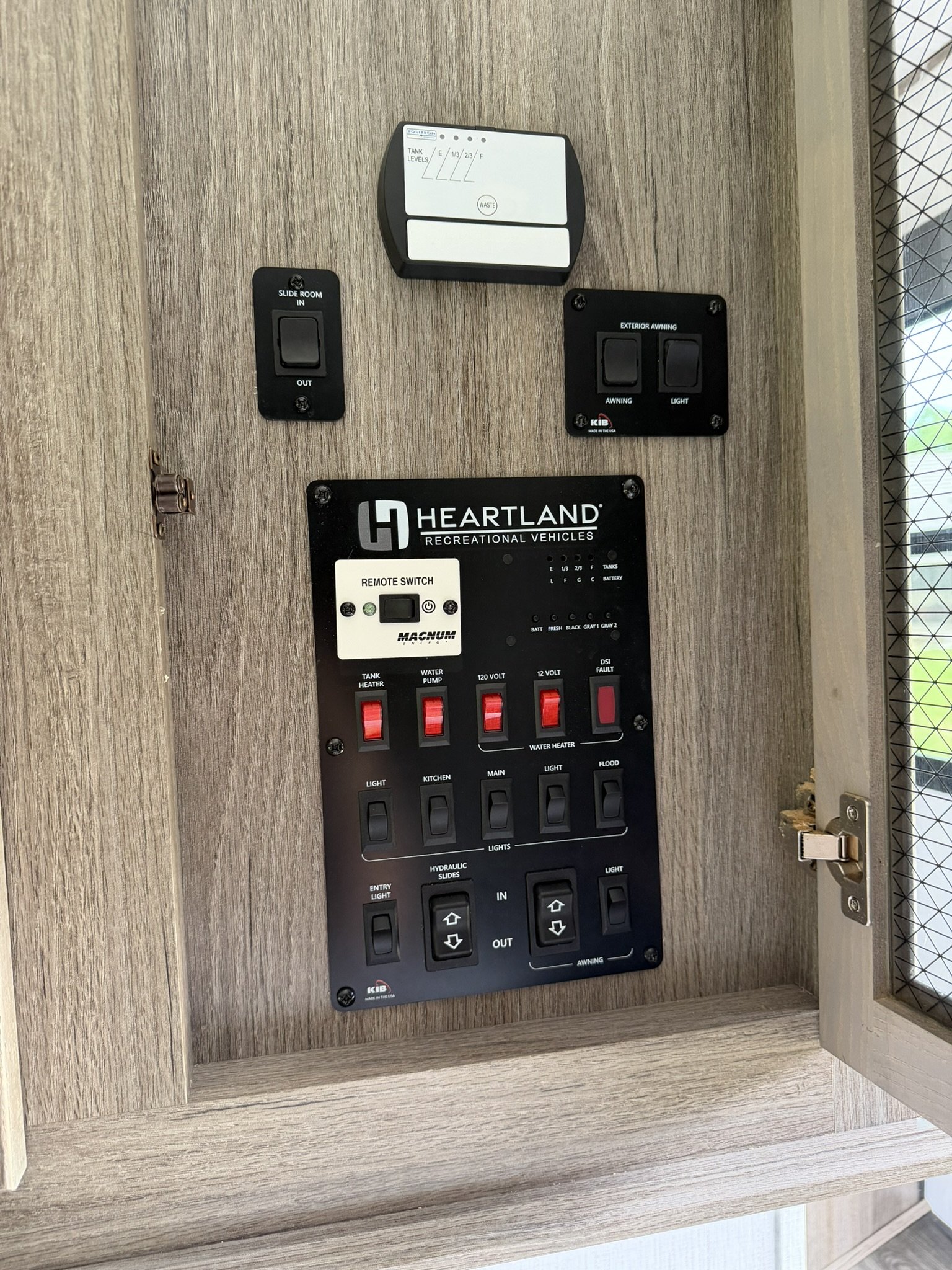 View of control panel and switches inside a recreational vehicle, including switches for slide room, awning, water pump, water heater, lighting, and entry light, mounted on a wood-paneled wall.