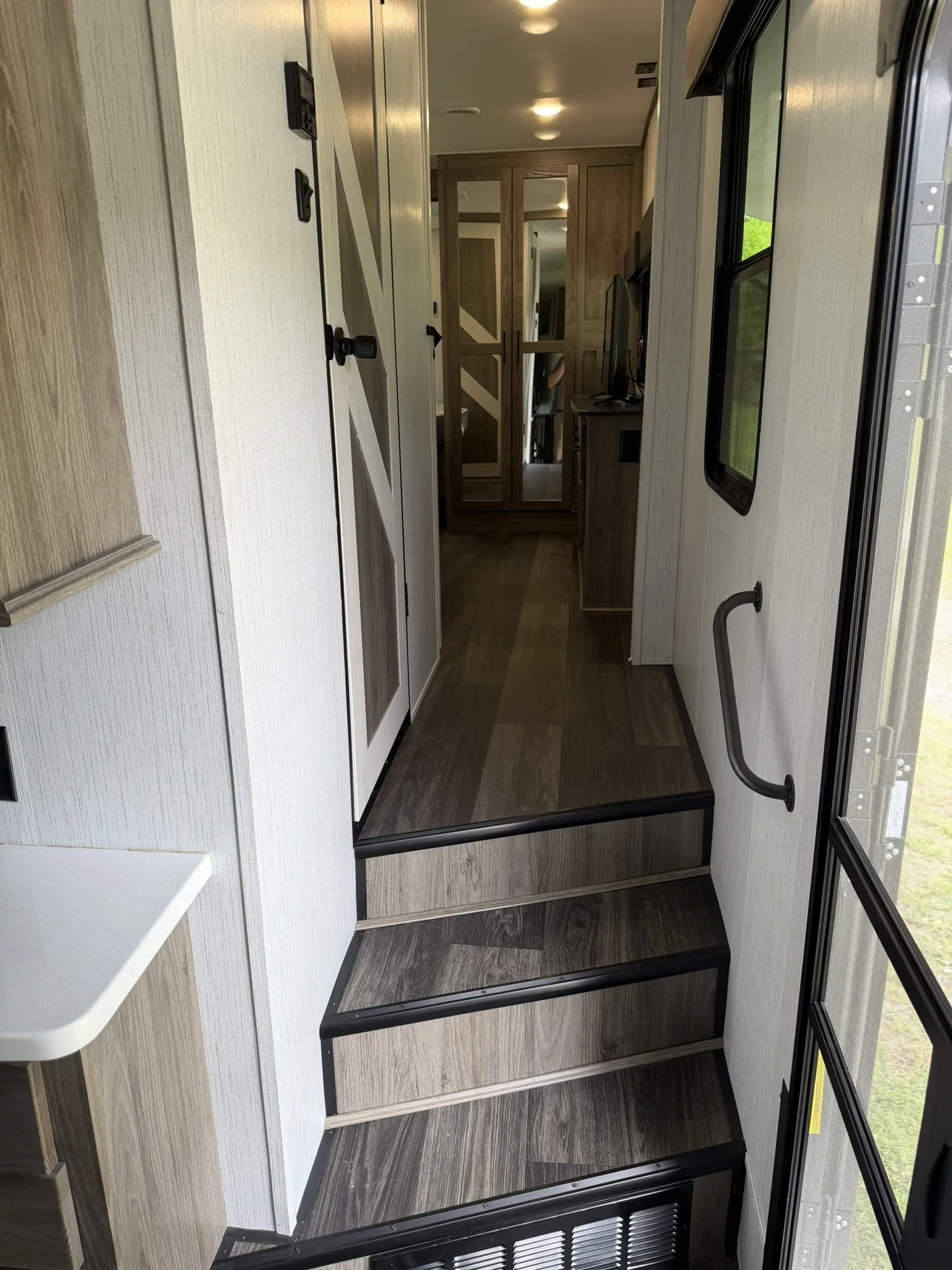 This image shows the interior of an RV, with a small set of three black steps leading into the living area, which has wood accents, a mirror cabinet, and a door to what appears to be a bathroom or bedroom.