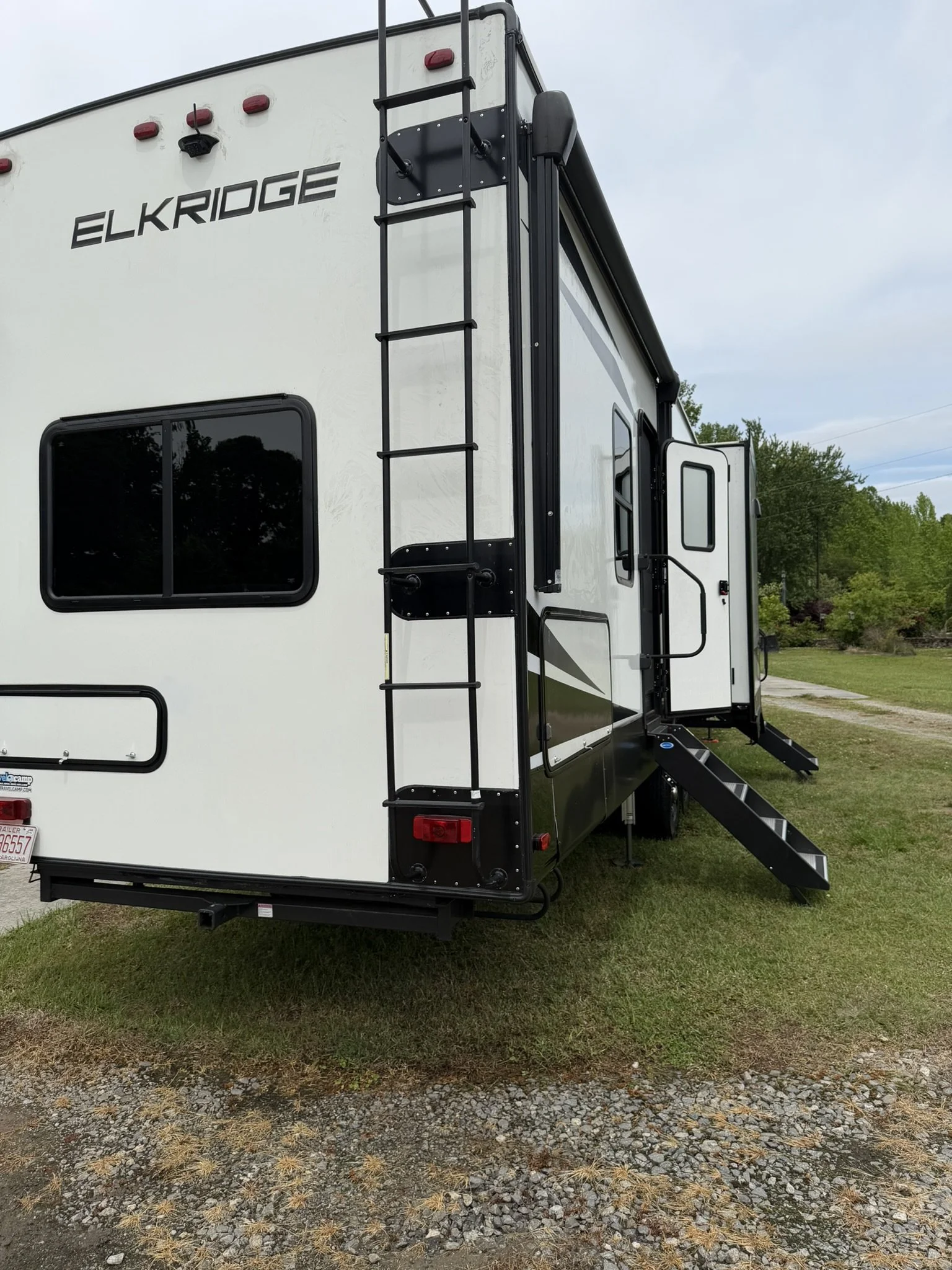 Rear view of a whitehorse camper trailer with black accents and a black ladder attached to the back, set on a grassy area with trees and a cloudy sky in the background.