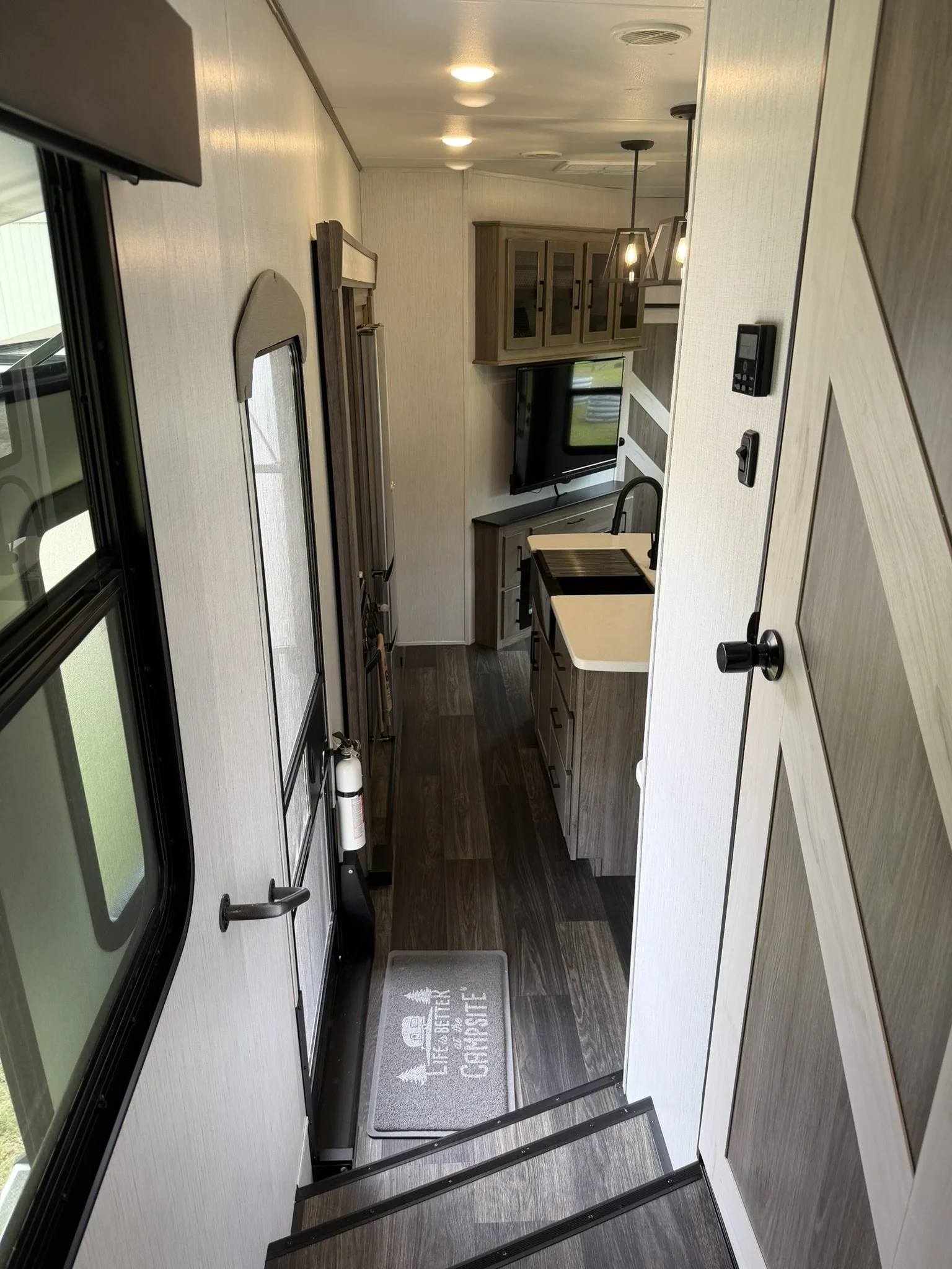 Interior view of a compact RV kitchen and living area with a small sink, wood cabinets, a mounted TV, and a door on the left, with a door mat on the dark wood flooring.