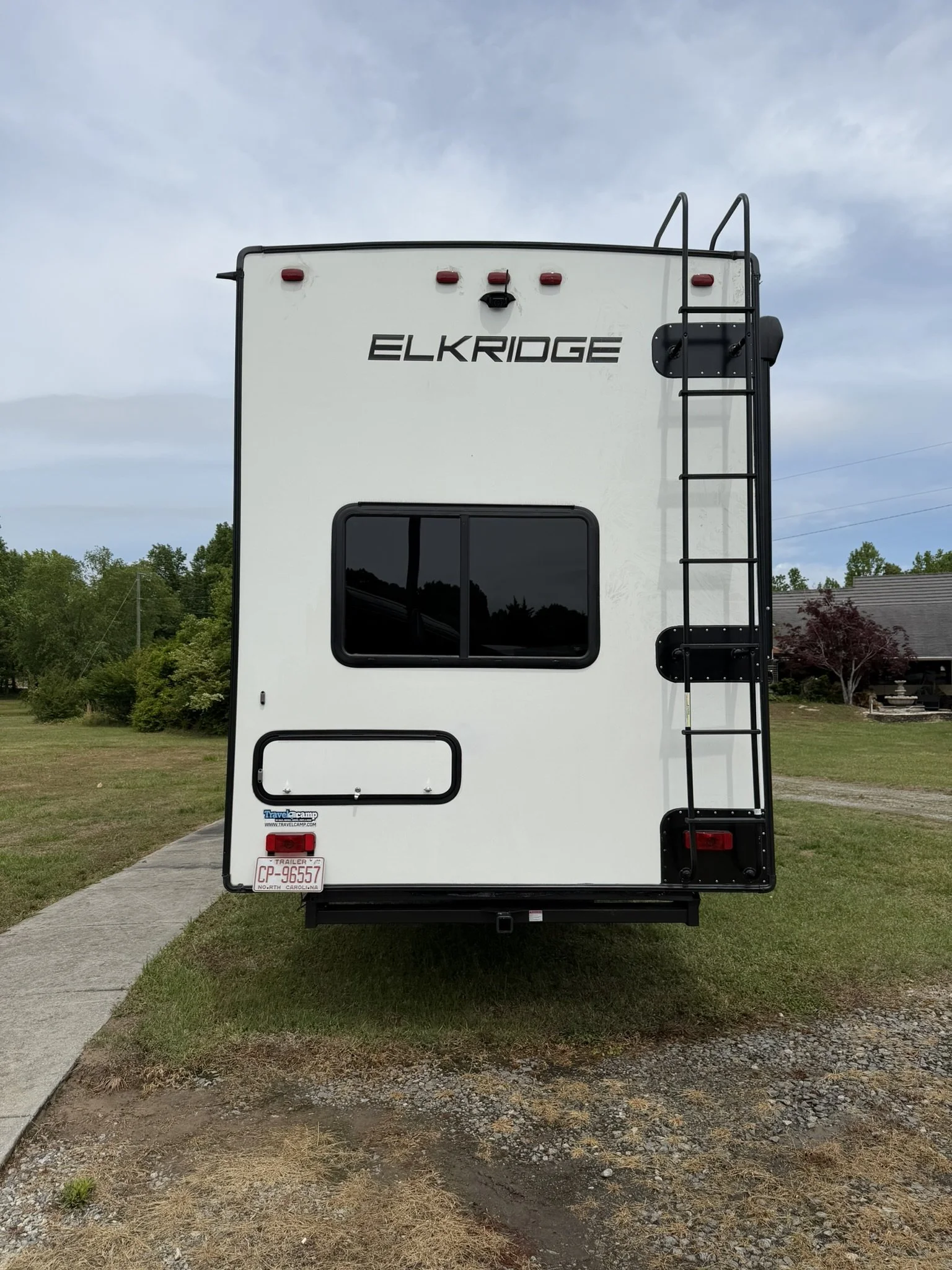 Rear view of a white travel trailer with the brand name ELKRIDGE, a black ladder on the right side, tinted window, and red brake lights, situated on a grassy area with trees and a house in the background under a cloudy sky.
