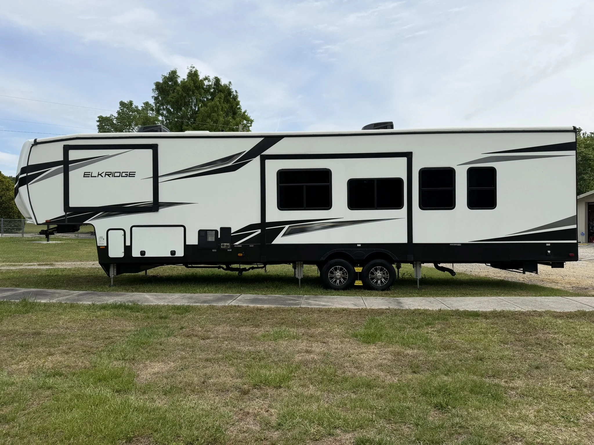 A white travel trailer with black and gray graphics parked on a grassy area, with trees and a partly cloudy sky in the background.