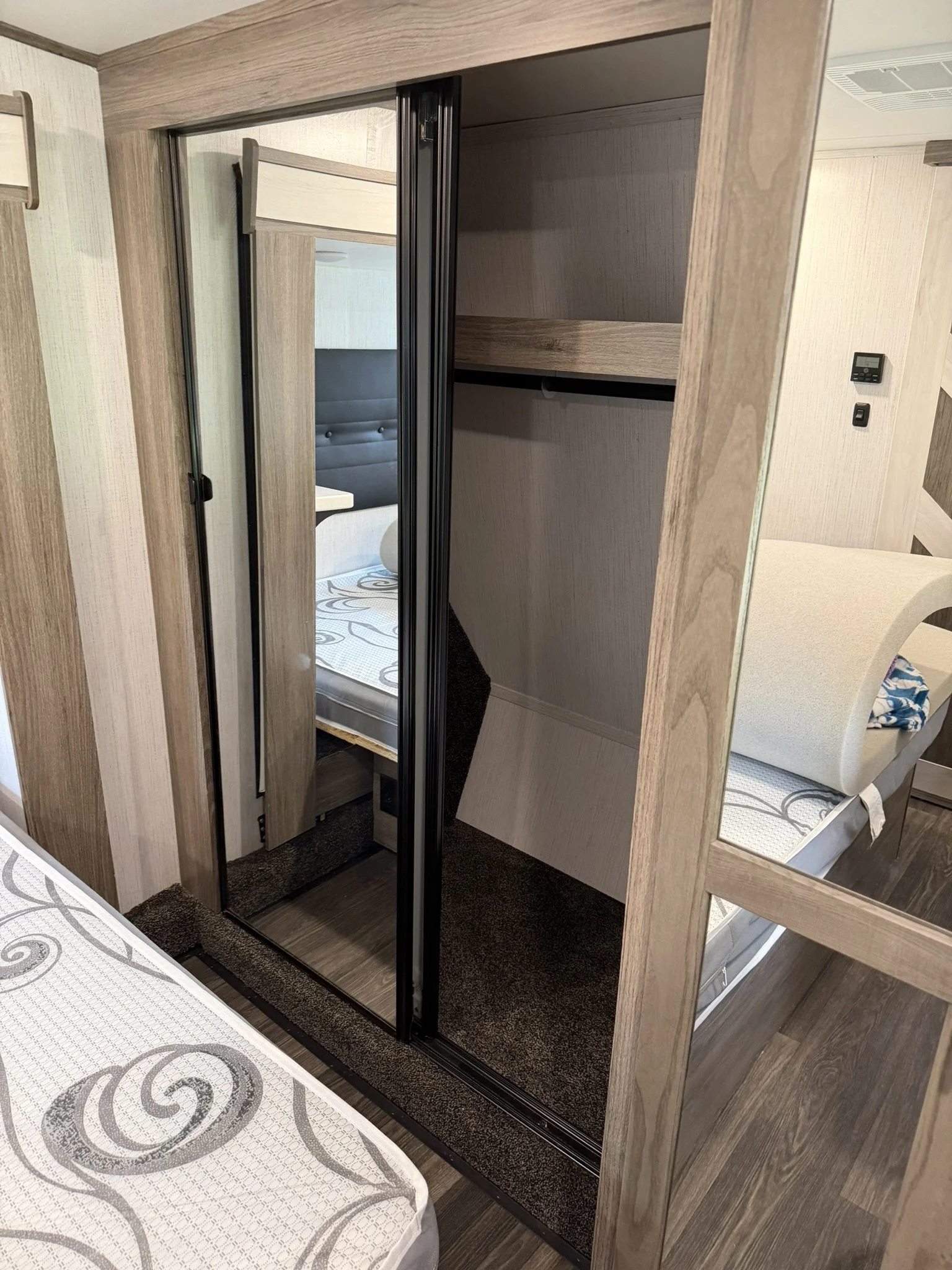 A sliding mirror closet door inside a wooden frame, with a bedroom visible through the open door showing a bed with a cushioned headboard, and a mattress with a decorative pattern on the sheets.