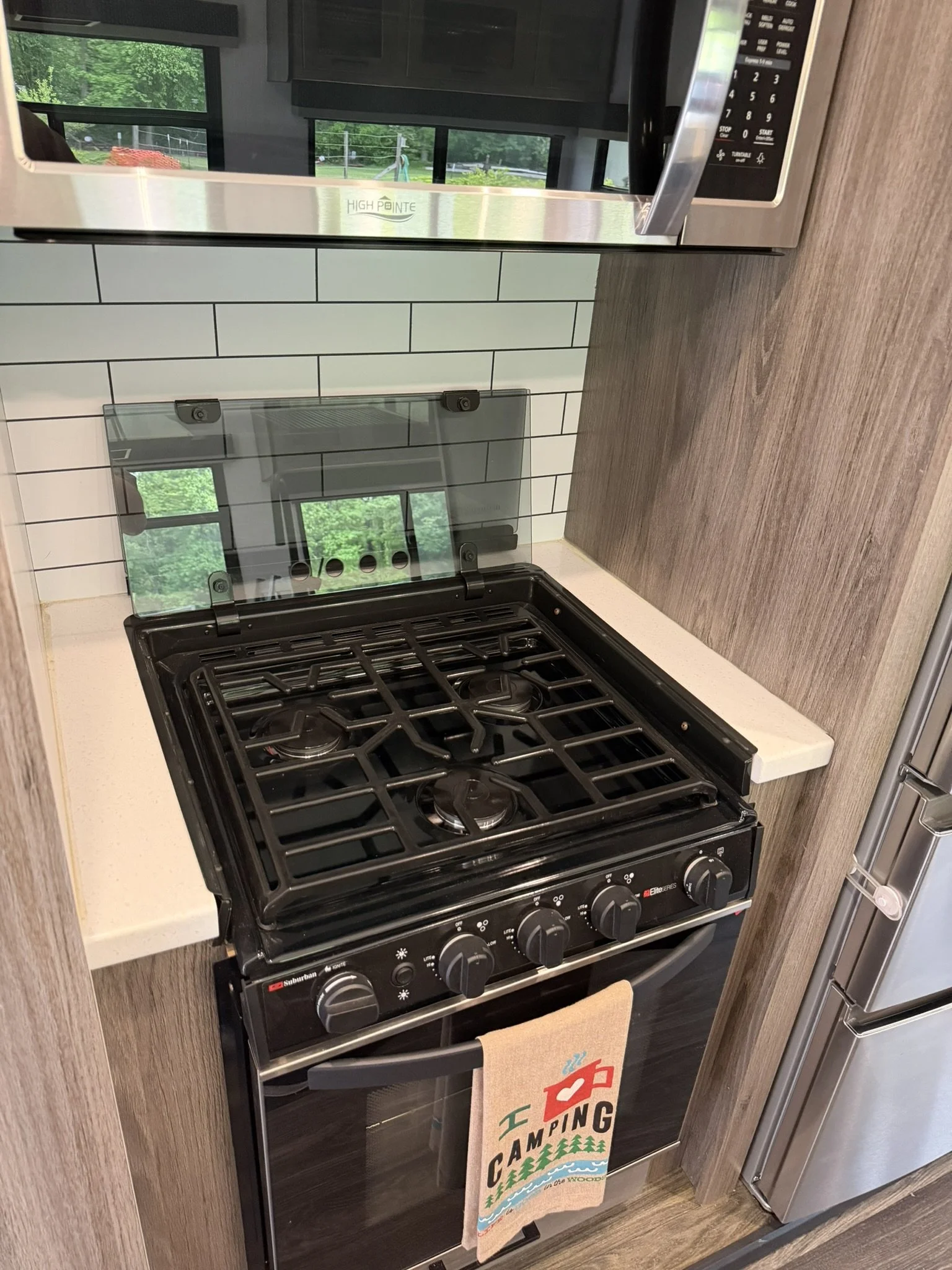 A kitchen with a black gas stove and oven, a beige dish towel hanging from the oven handle with a camping-themed design, a wooden countertop, a white subway tile backsplash, a microwave above the stove with a reflection of windows and greenery outside, and a refrigerator on the right side.