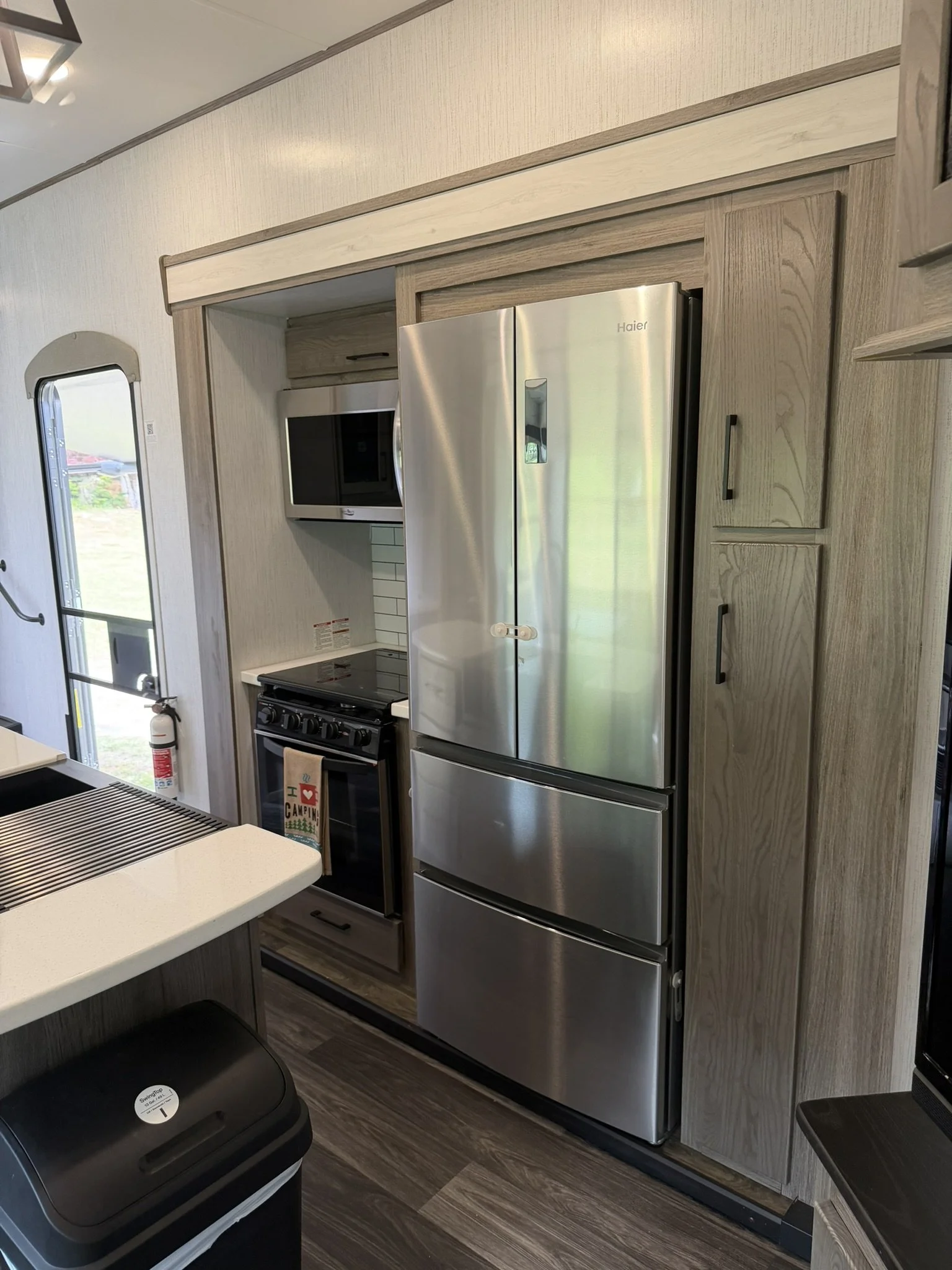 Kitchen area with a stainless steel Haier refrigerator, a black stove with oven, white countertop, wall-mounted microwave, window, and wooden cabinets.
