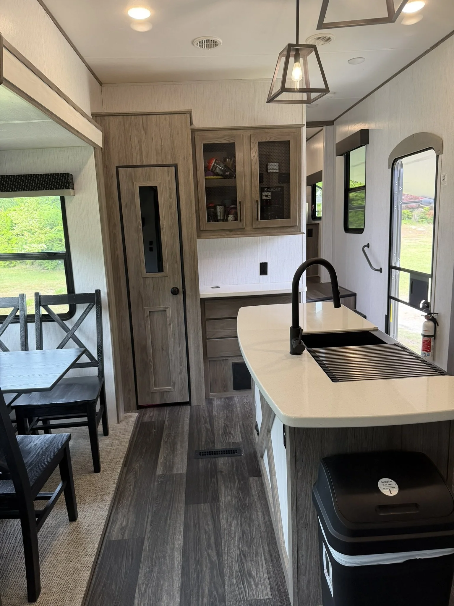 Kitchen area with a countertop, sink, black faucet, and black trash bin, surrounded by wooden cabinets, windows with green scenery outside, and a ceiling light fixture.