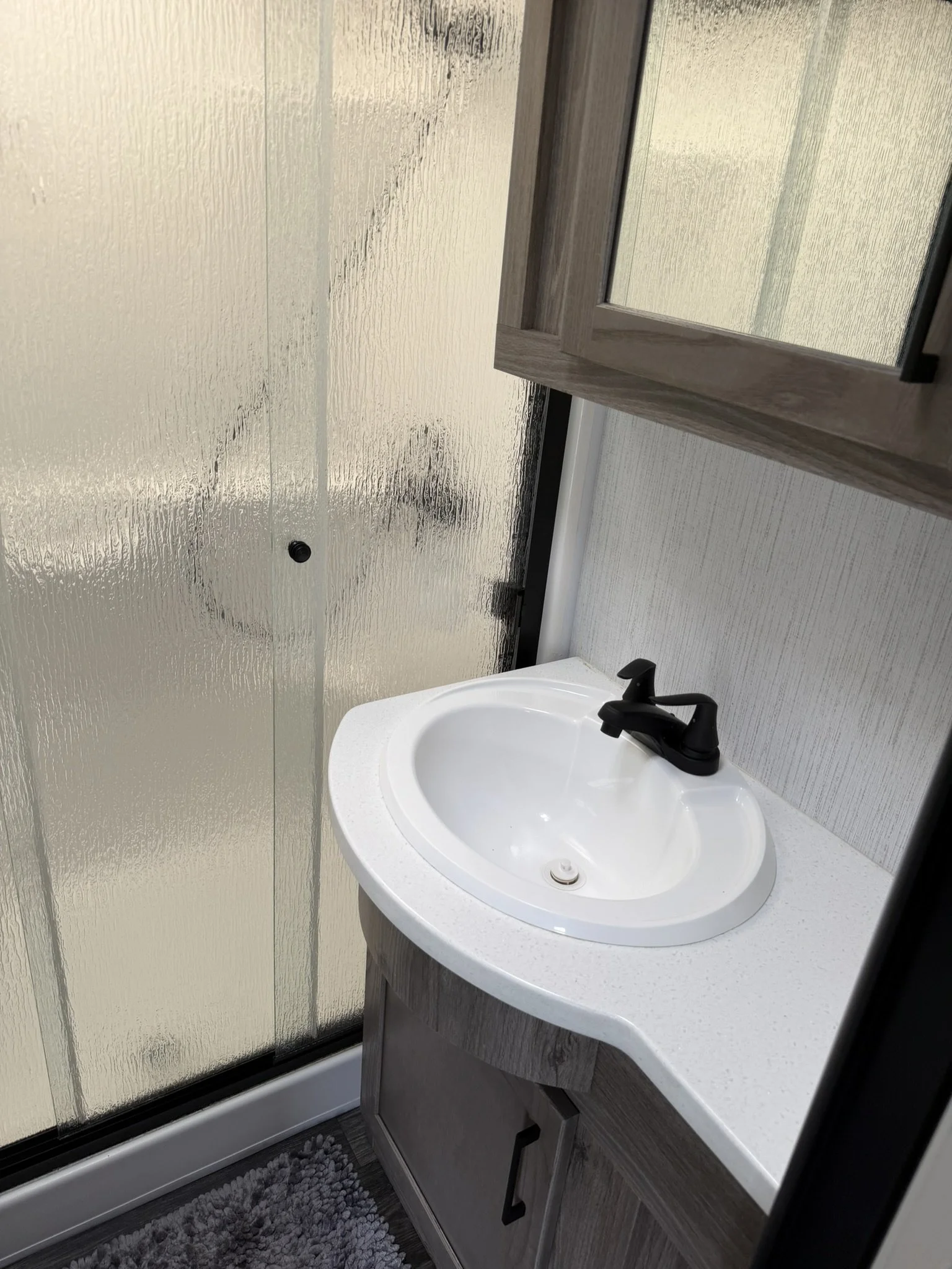 A small white bathroom sink with a black faucet, mounted on a wooden cabinet with black handle, positioned next to a frosted glass shower door and a textured wall. A gray shaggy rug is on the floor nearby.
