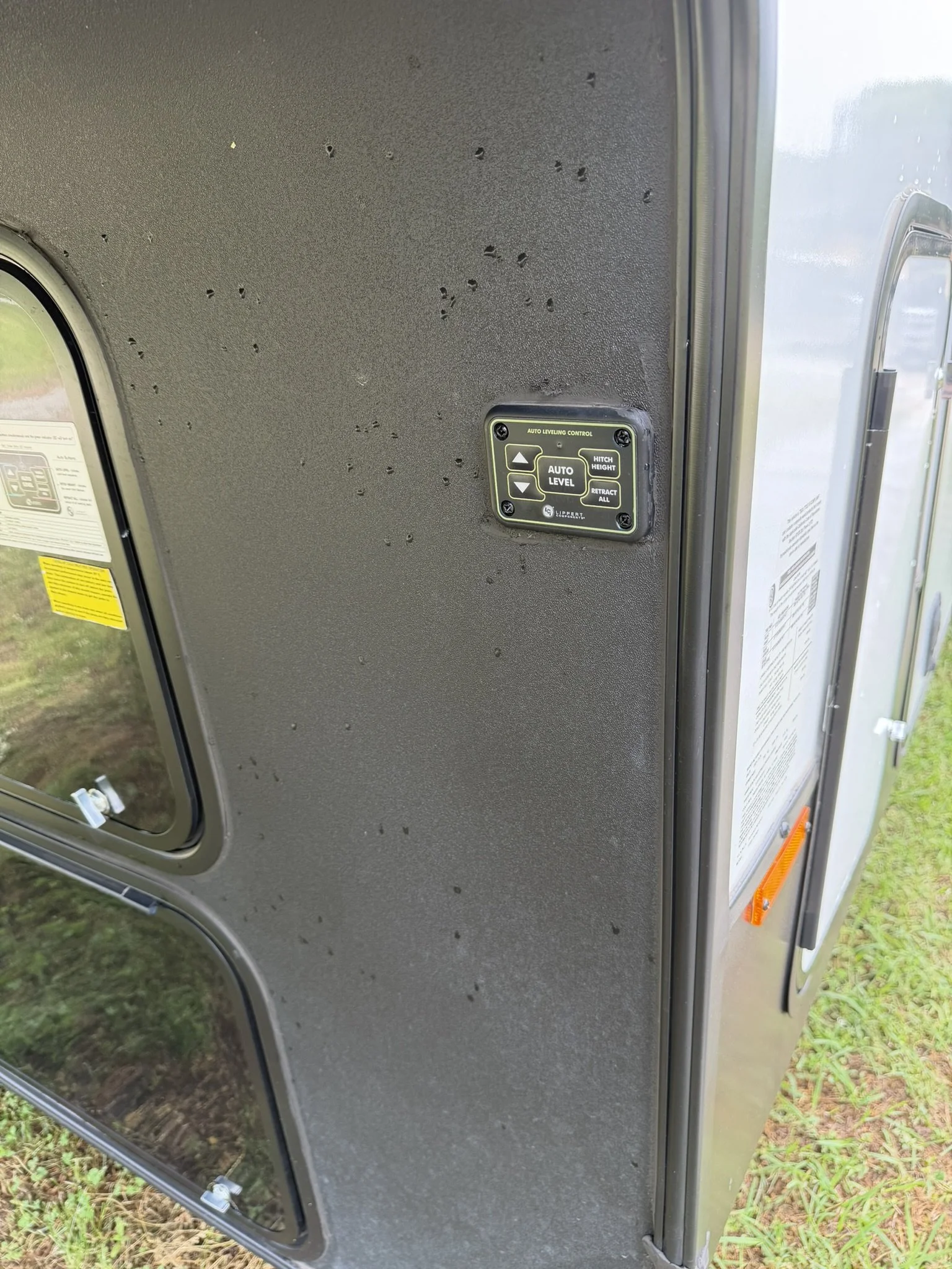 Close-up of a gray trailer wall with a small control panel labeled 'Auto Leveling Control' featuring buttons for adjusting hitch height, retracing all, and auto level, seen outdoors with grass in the background.