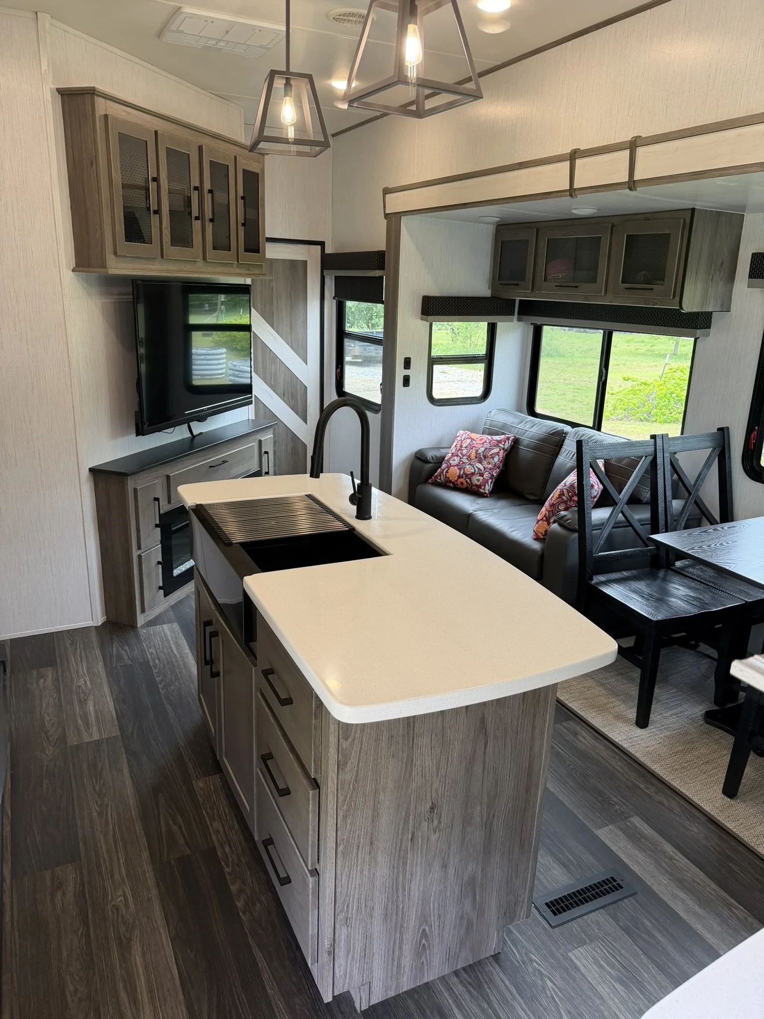 Inside view of a modern RV or tiny home with a kitchen island, a seating area with a gray sofa and colorful cushions, and overhead cabinets with glass doors. There are windows with black trim, and a dining table with black chairs to the right. The walls are light-colored and the flooring is dark wood. A pendant light fixture hangs from the ceiling.