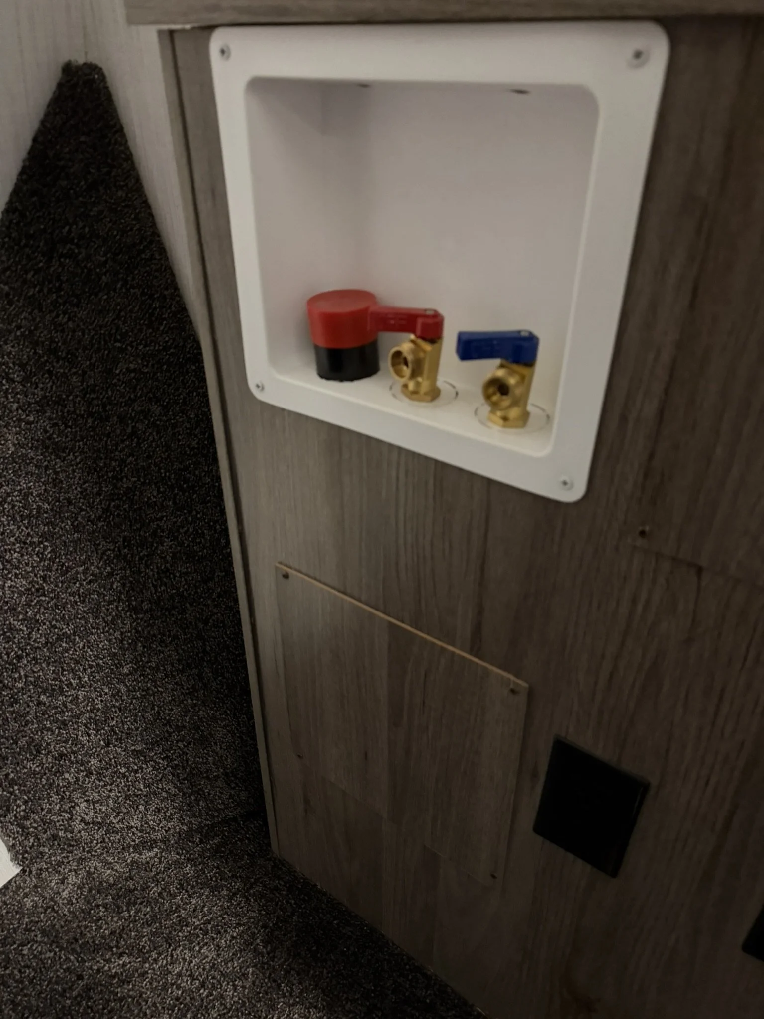 An electrical outlet with red and blue shut-off valves on a wooden surface, with black vent and gray carpeting nearby.