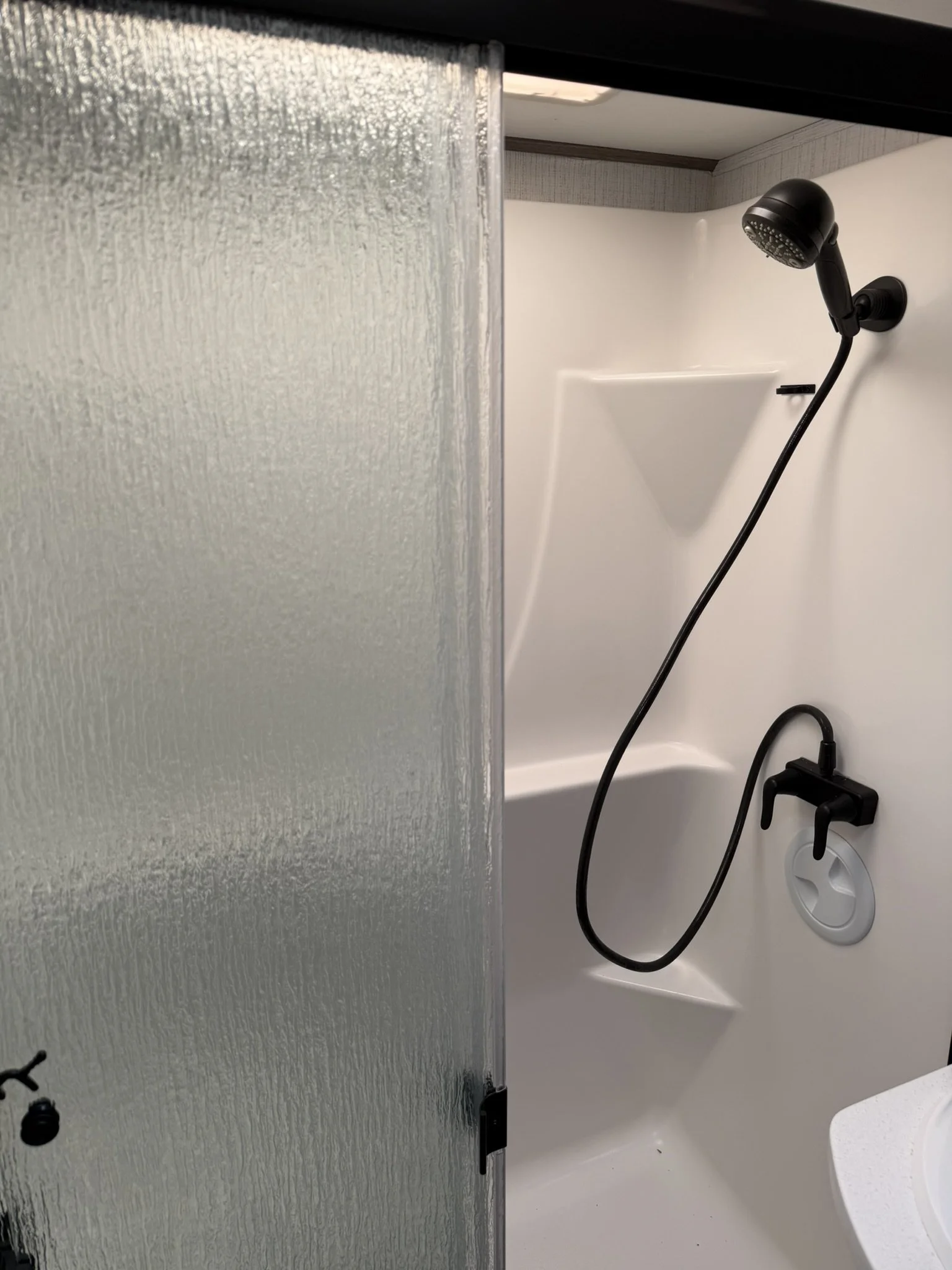 A shower stall with a black handheld showerhead attached to a flexible hose, a small soap ledge, and a textured frosted glass door, with a white plastic drain cover at the bottom.