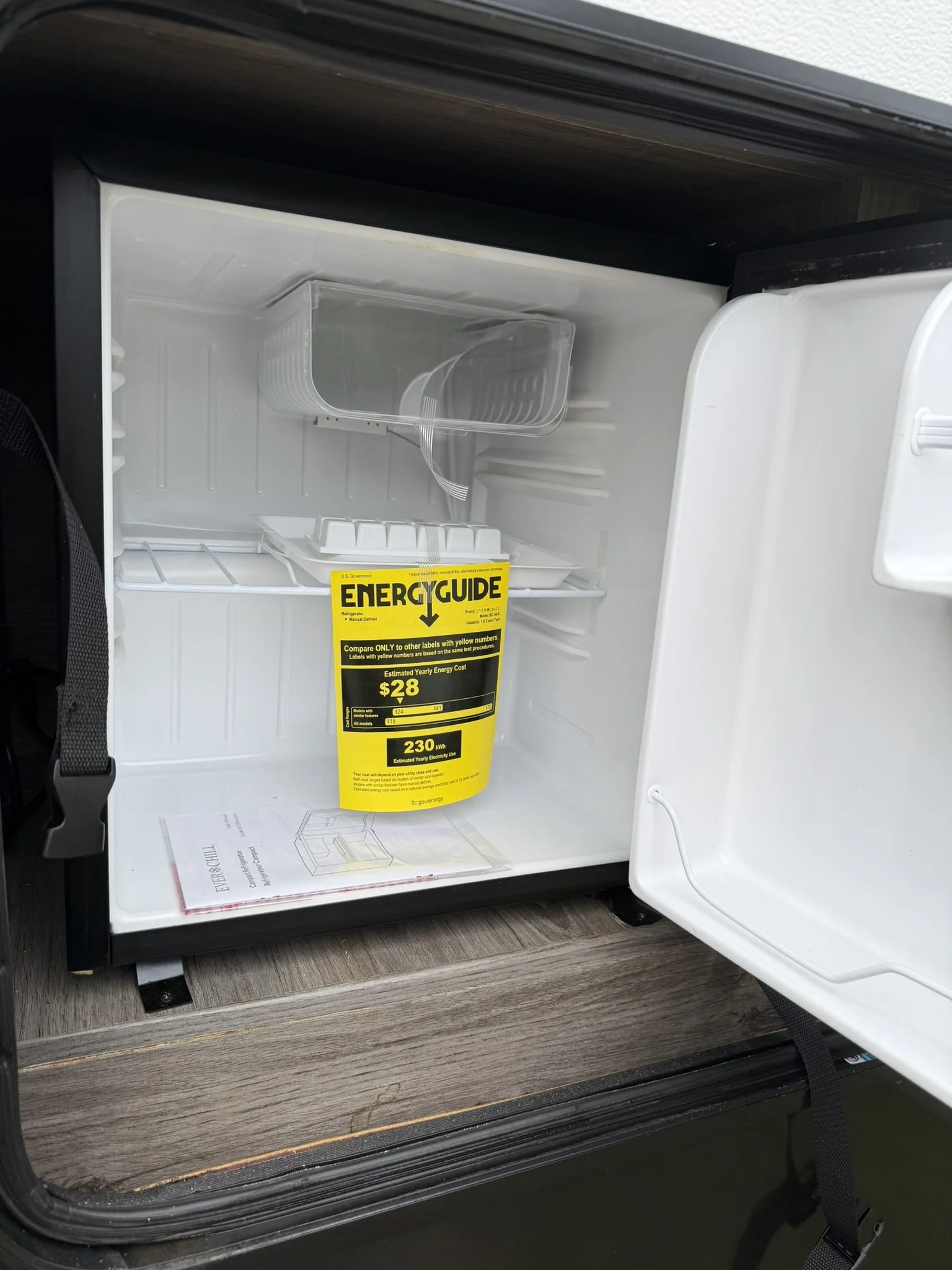 Empty mini refrigerator with a yellow energy guide label on the door and a diagram manual inside, set on a wooden surface.