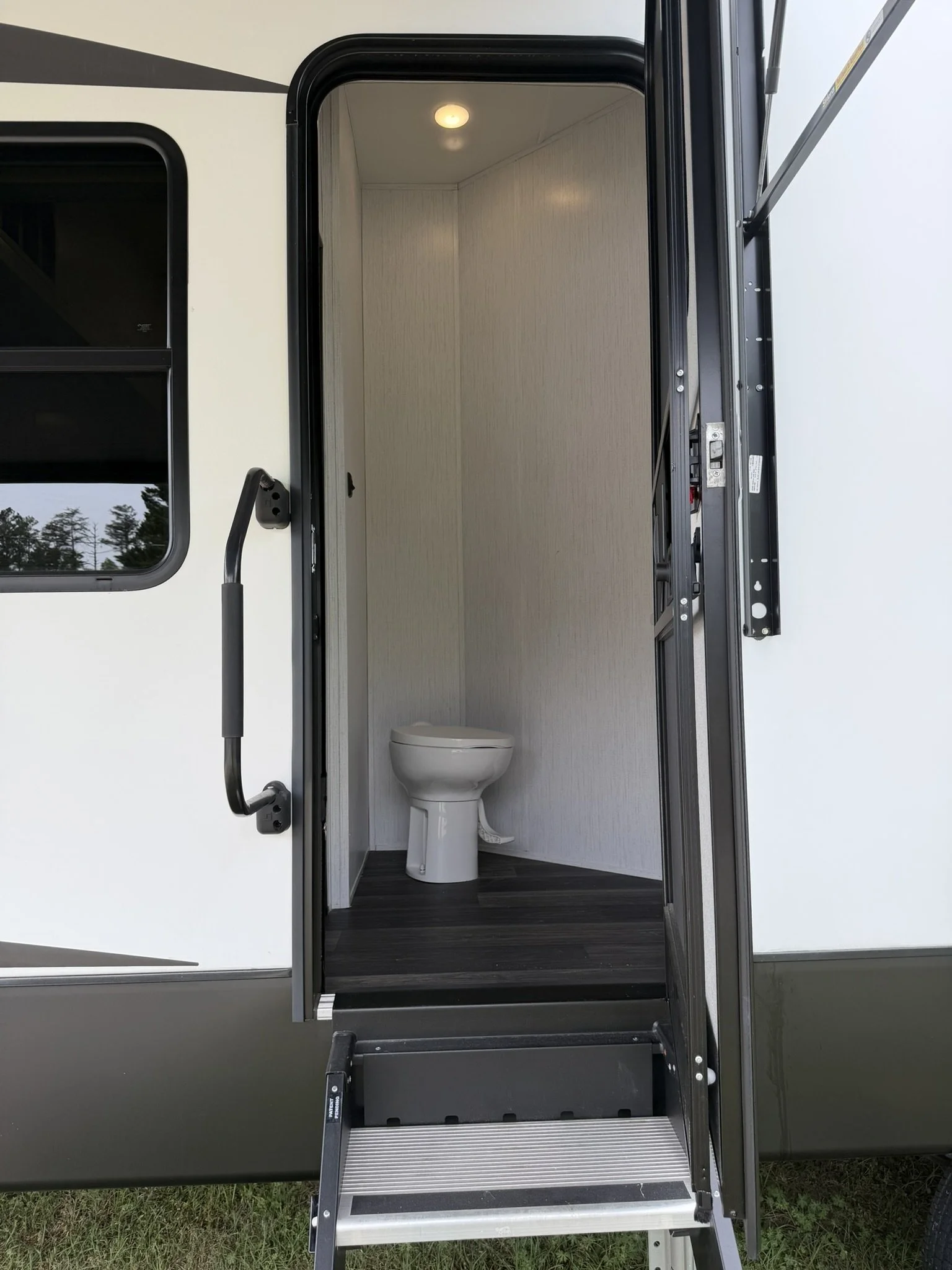 Exterior view of a portable restroom with open door revealing a toilet inside, set on grass.