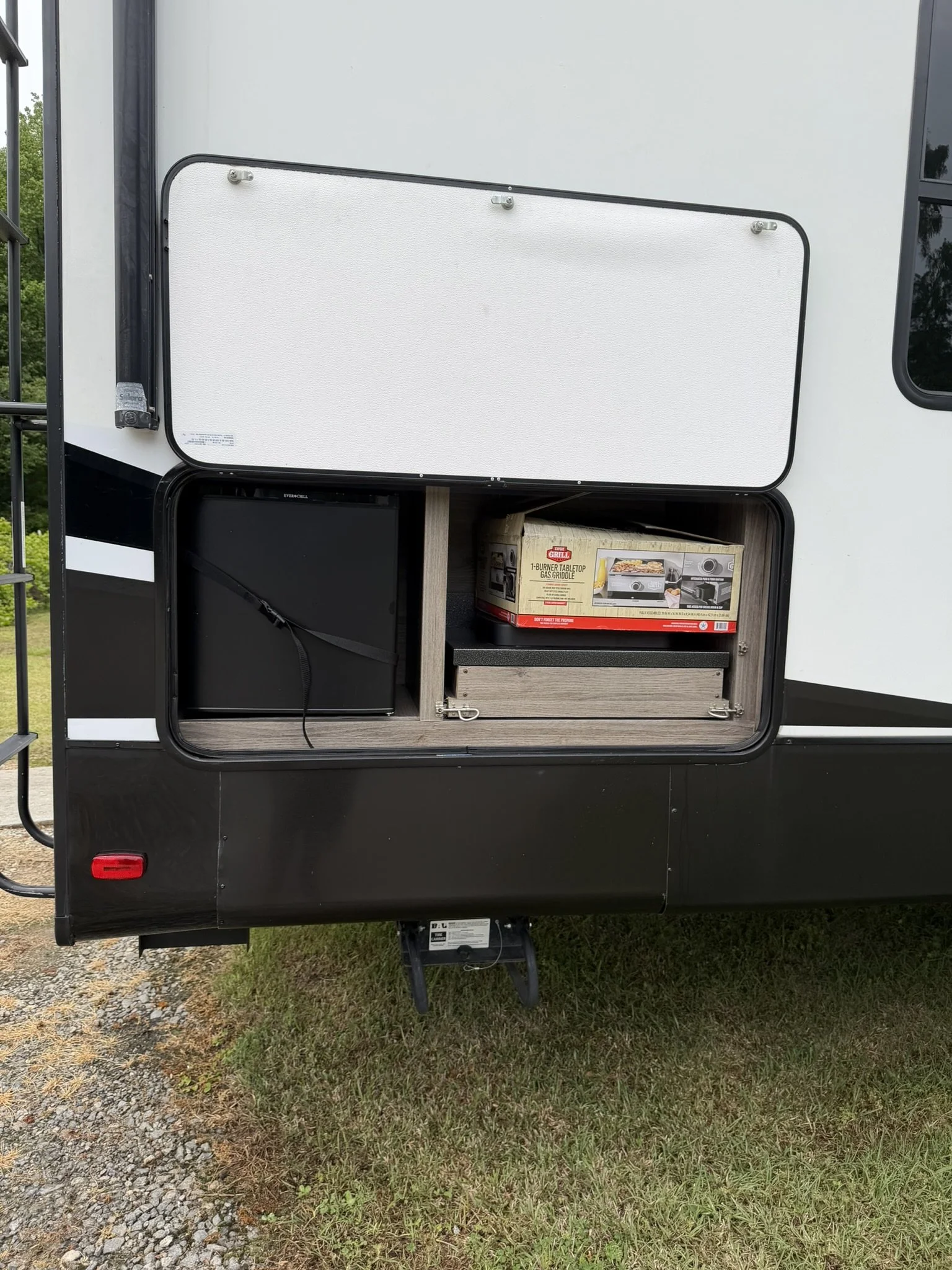 The side view of a recreational vehicle with an outdoor storage compartment containing a boxed gas grill and a black mini refrigerator.
