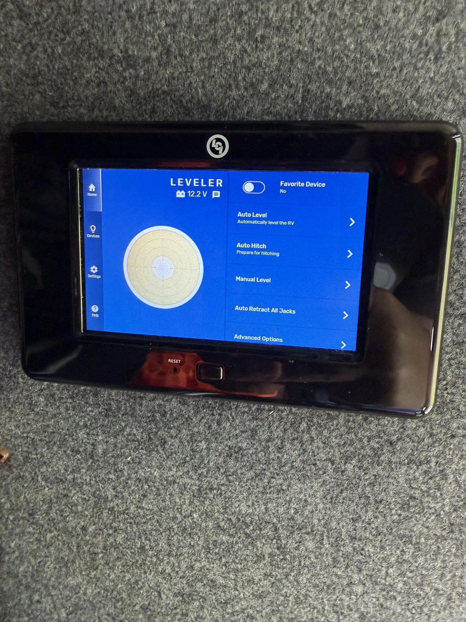 A digital level display screen showing a blue interface with a circular level indicator, menu options for auto level, auto hitch, manual level, and advanced options, and a toggle for favorite device, set against a gray textured surface.