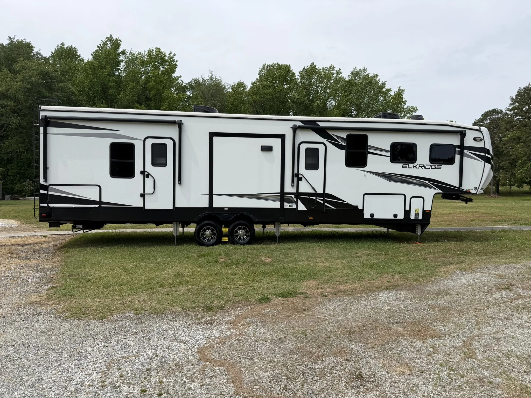 A white travel trailer parked on grass with green trees in the background under a cloudy sky.