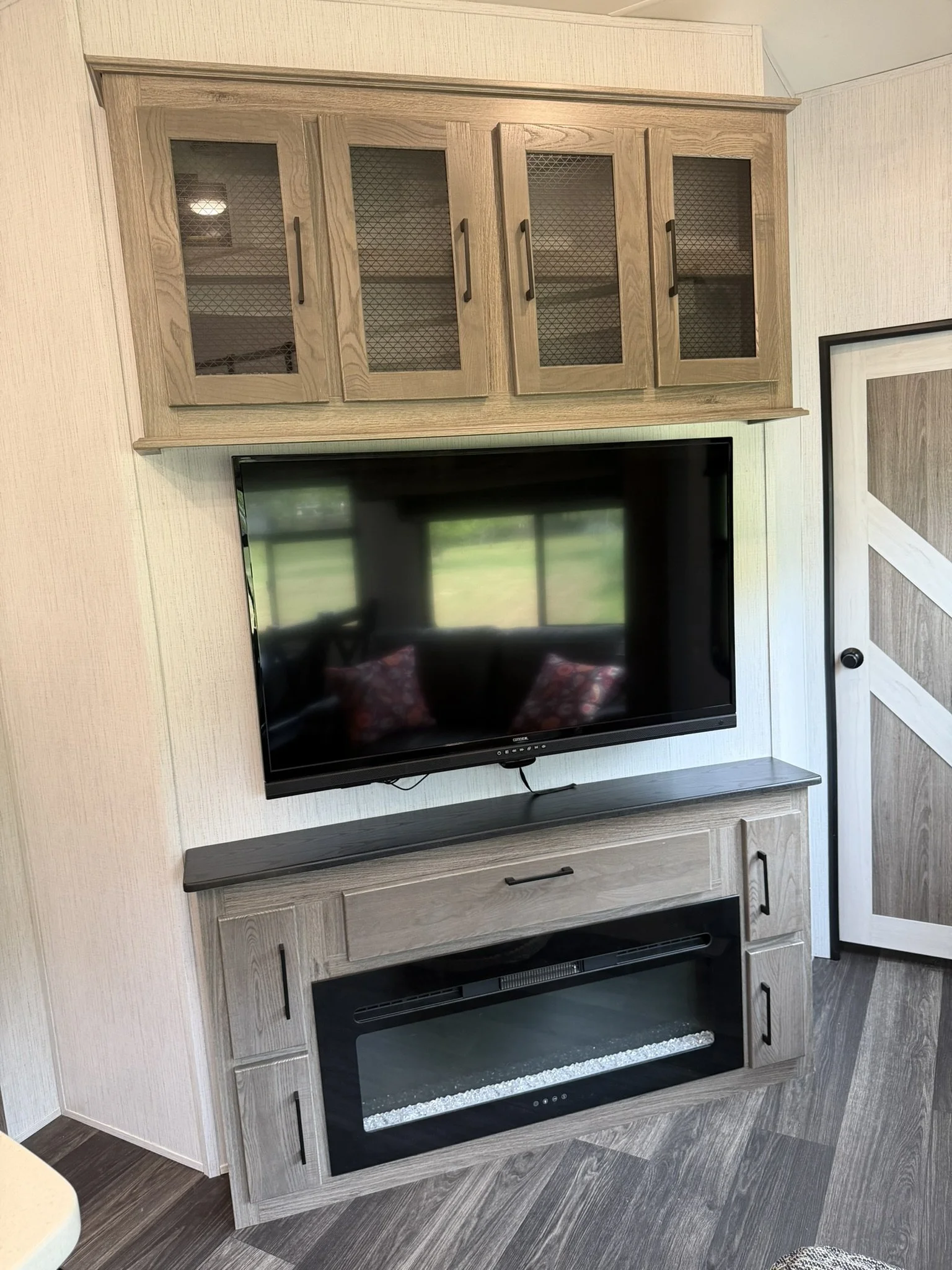 Living room media wall with a large flat-screen TV mounted above an electric fireplace, and a wooden cabinet with glass doors and black handles above the TV.