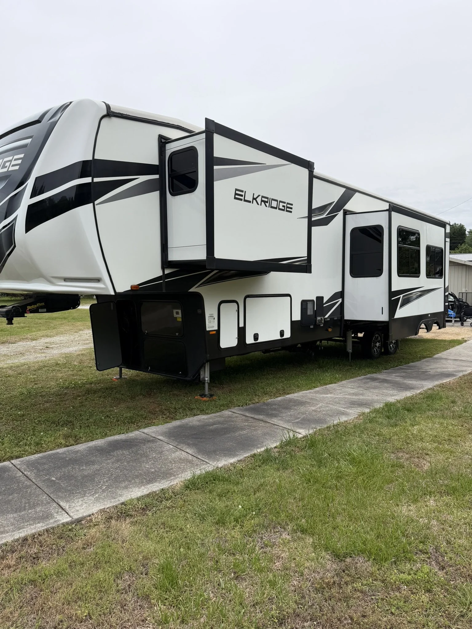 A white and black travel trailer with the brand name 'ELKRIDGE' on its side parked on grass with a sidewalk in front and a building in the background.