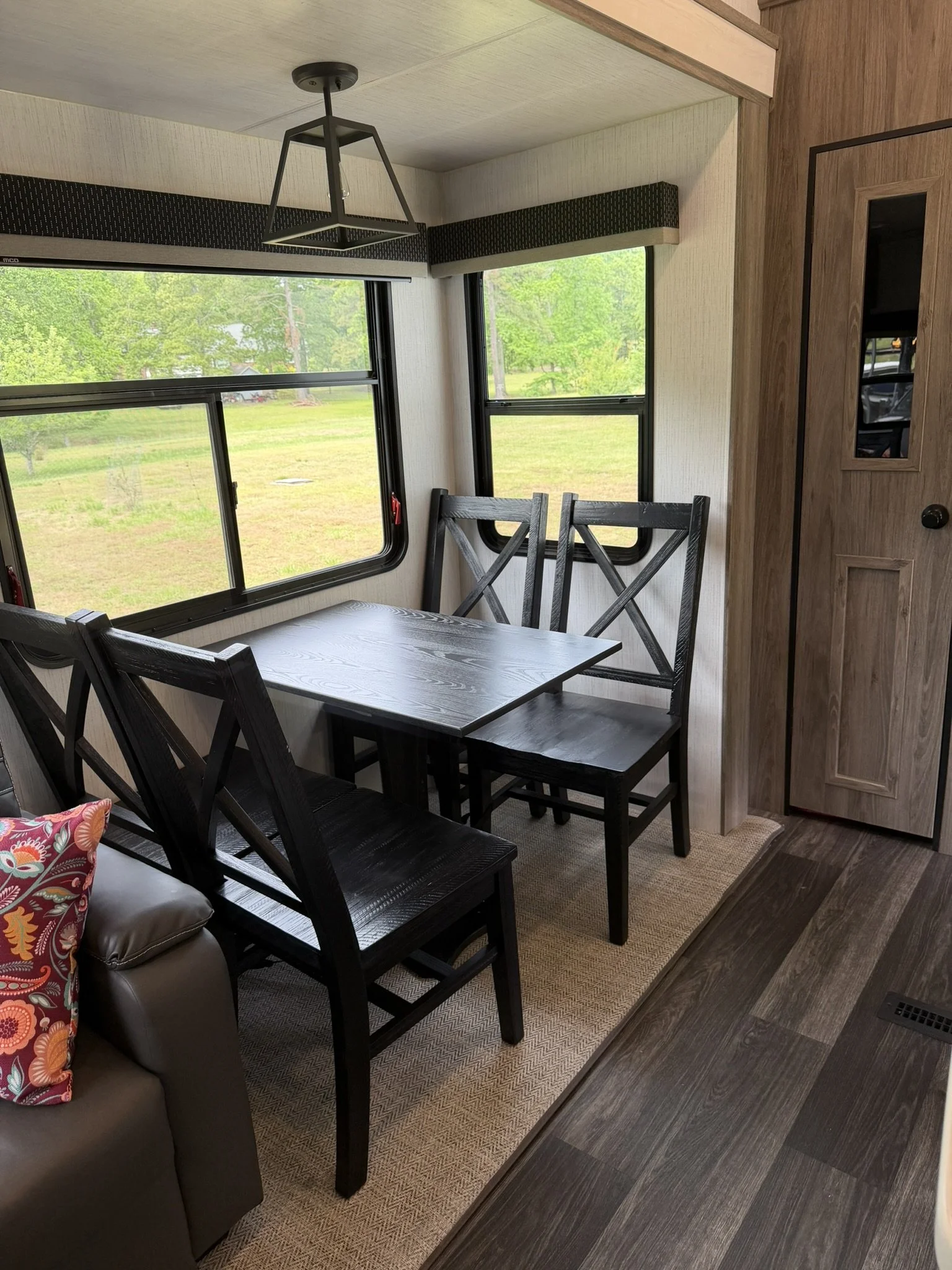 Interior view of a dining area in a camper or small home, showing a wooden table with four black chairs, a window with greenery outside, a ceiling light fixture, and a wooden door on the right.