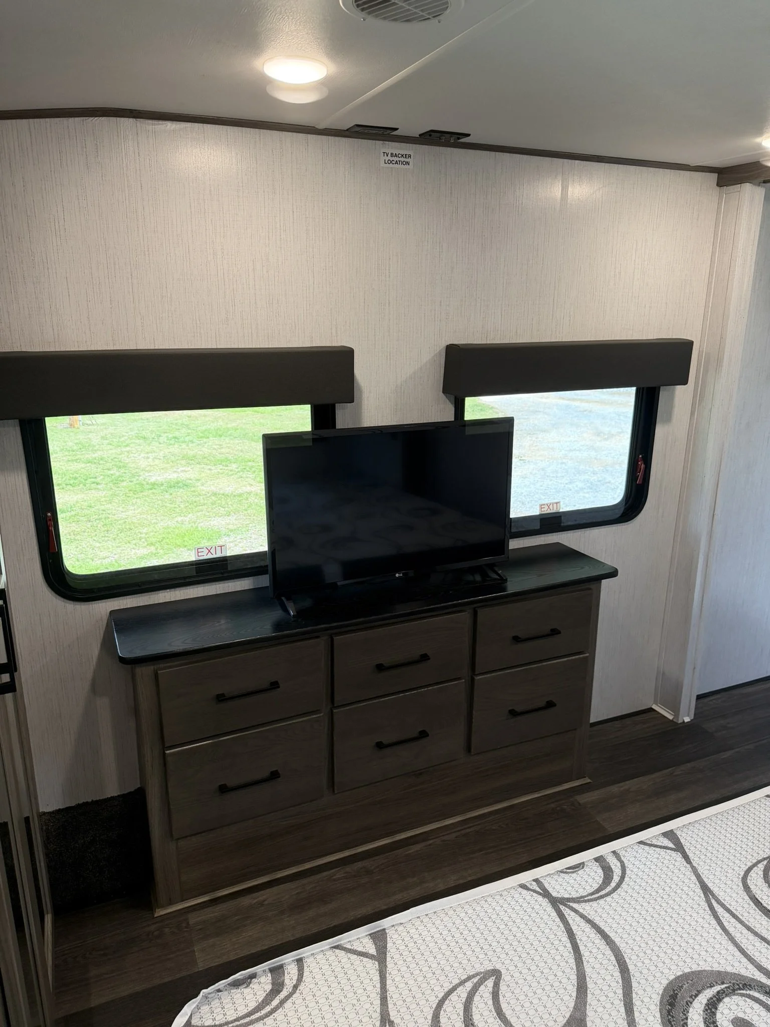 A section of a room with a wooden cabinet below two windows, a television on top, and a wall with a sign labeled 'TV BACKER LOCATION'. The room has ceiling lights and a patterned rug on the floor.