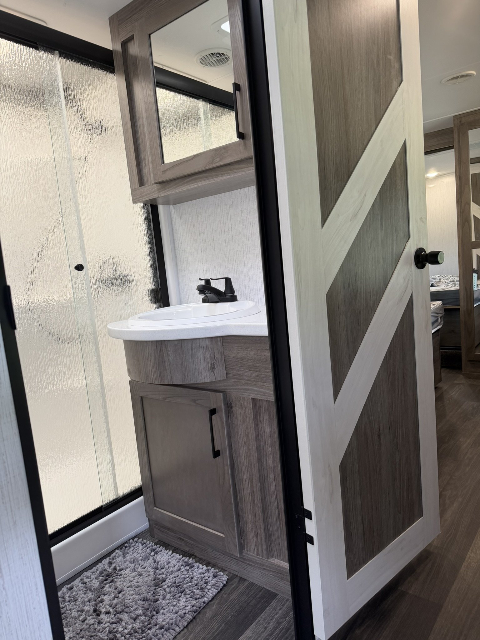 View of a bathroom with a small sink, wooden cabinets, a mirror, and a textured glass shower door.