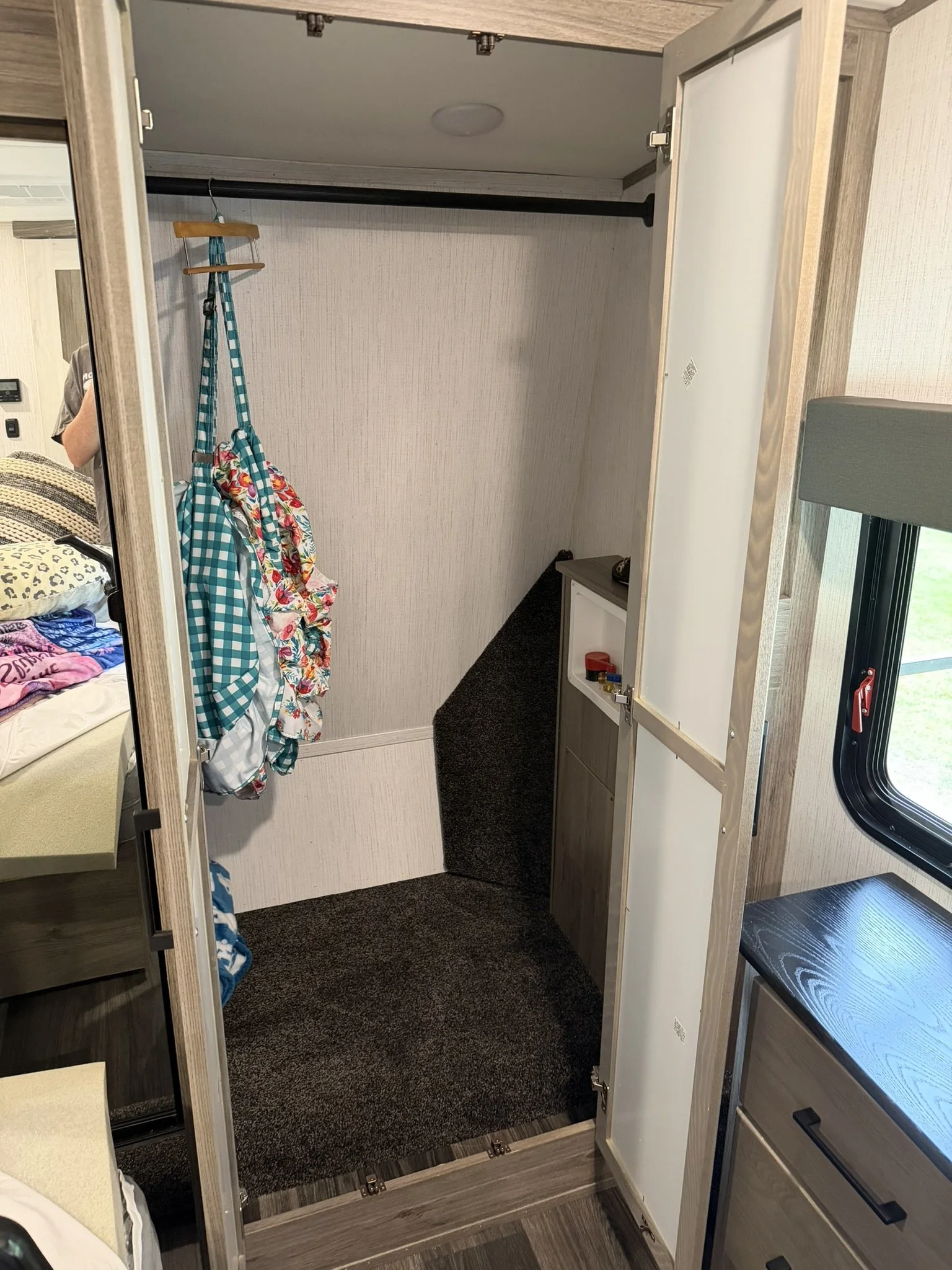 Open closet with a hanging bar and a few clothes hanging, a small cabinet with items inside, and a window on the right side, outside view not visible.