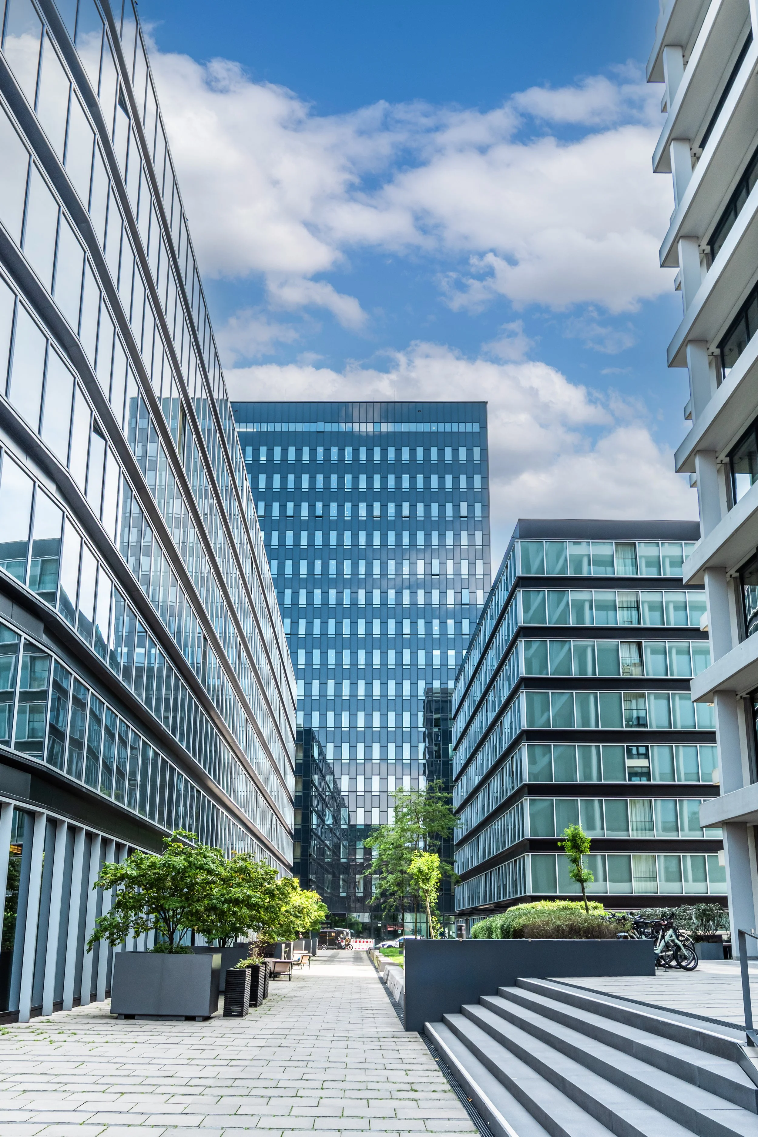 a cobblestone walkway with a short stairway on the right and plants and office buildings on either side and a larger office building in the background