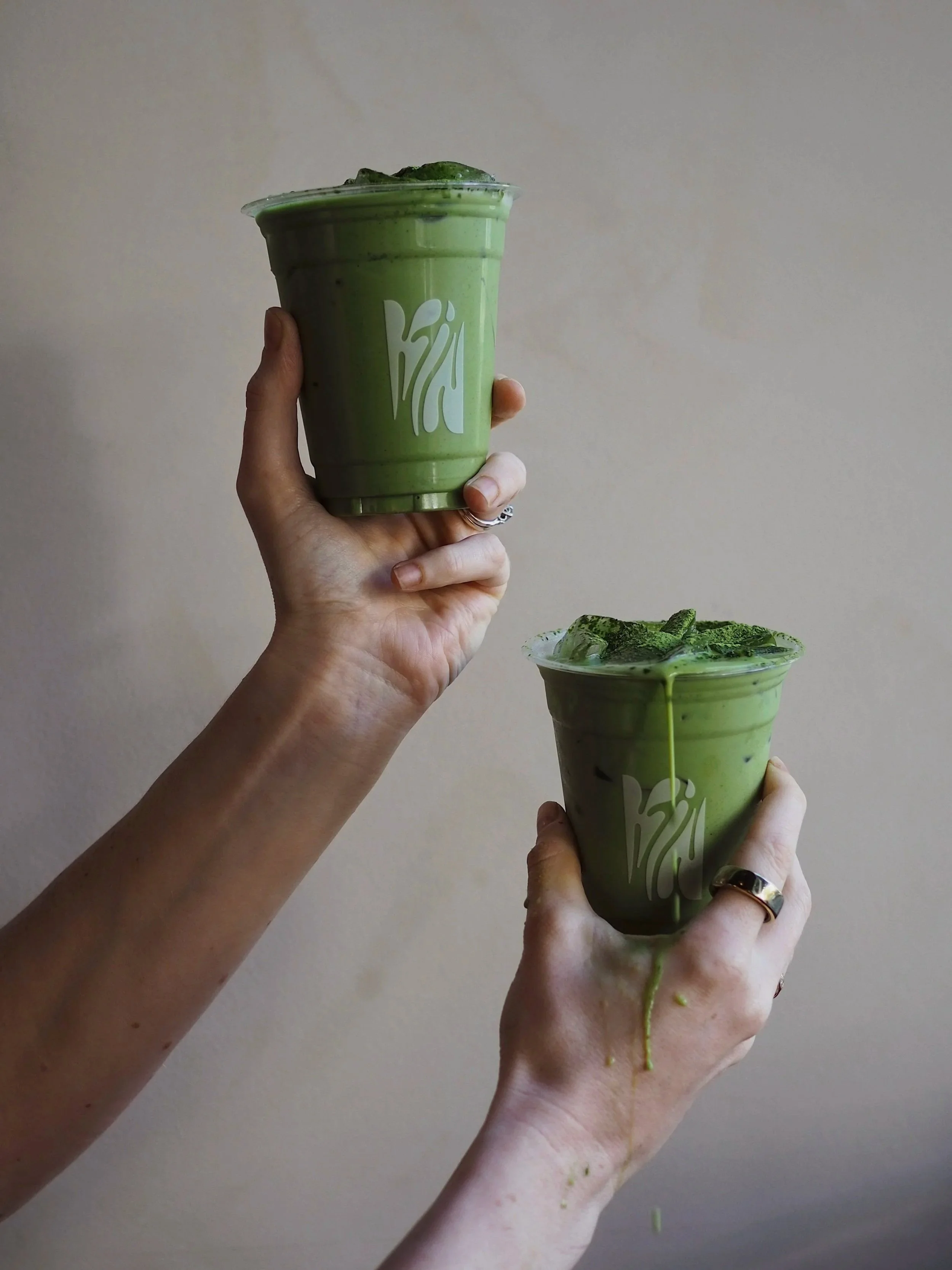 Two hands holding cups with green matcha drinks, one with ice and the other with a green whipped topping, against a plain beige background.