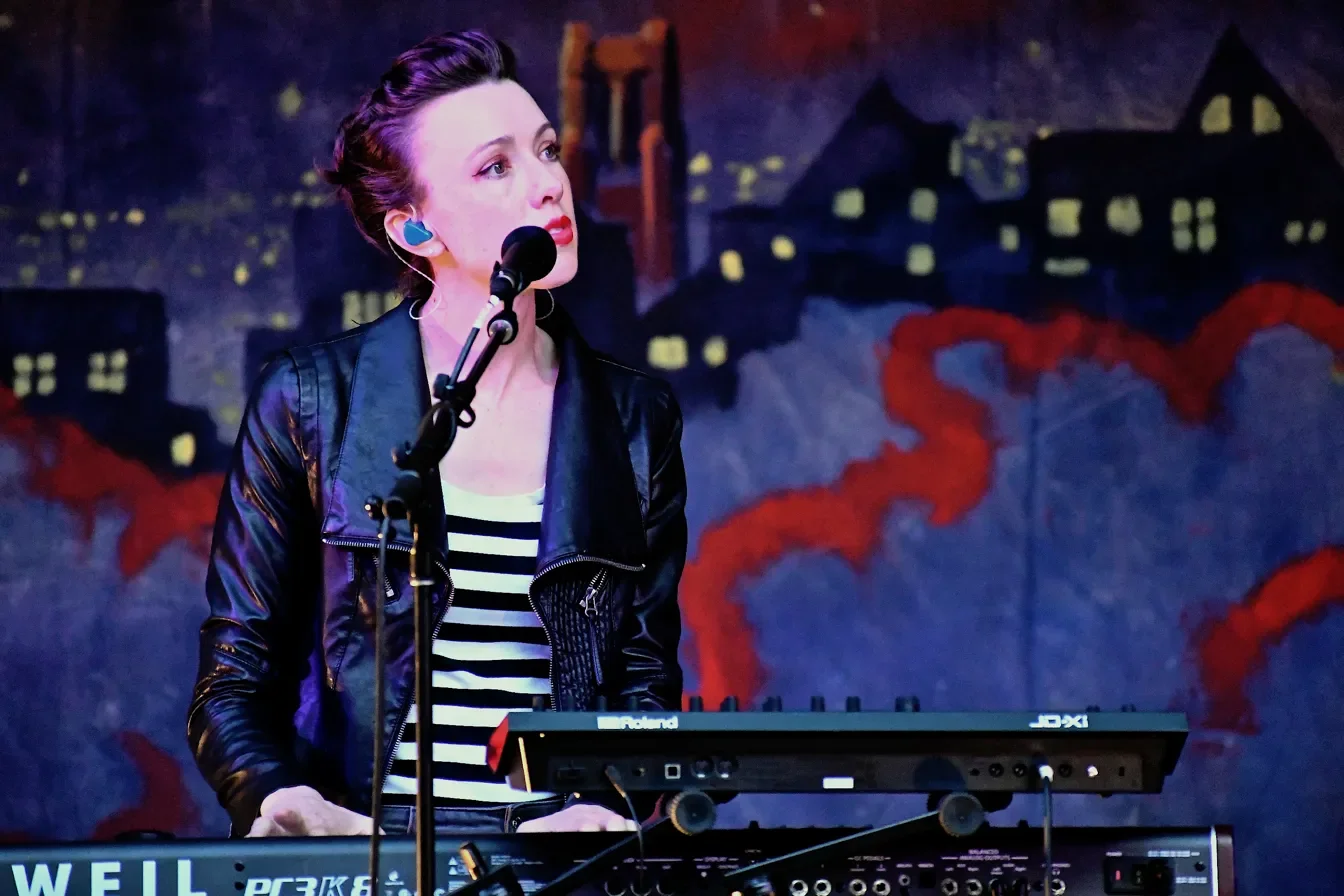 A woman with brown hair in an updo, wearing a black jacket and striped shirt, performs on stage using a keyboard with a microphone in front of her, against a painted cityscape backdrop.