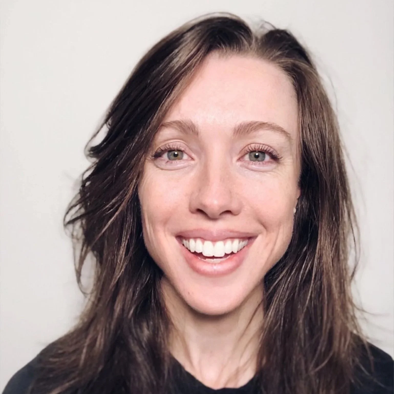Close-up of a smiling woman with long brown hair and green eyes, wearing a black top, against a plain background.