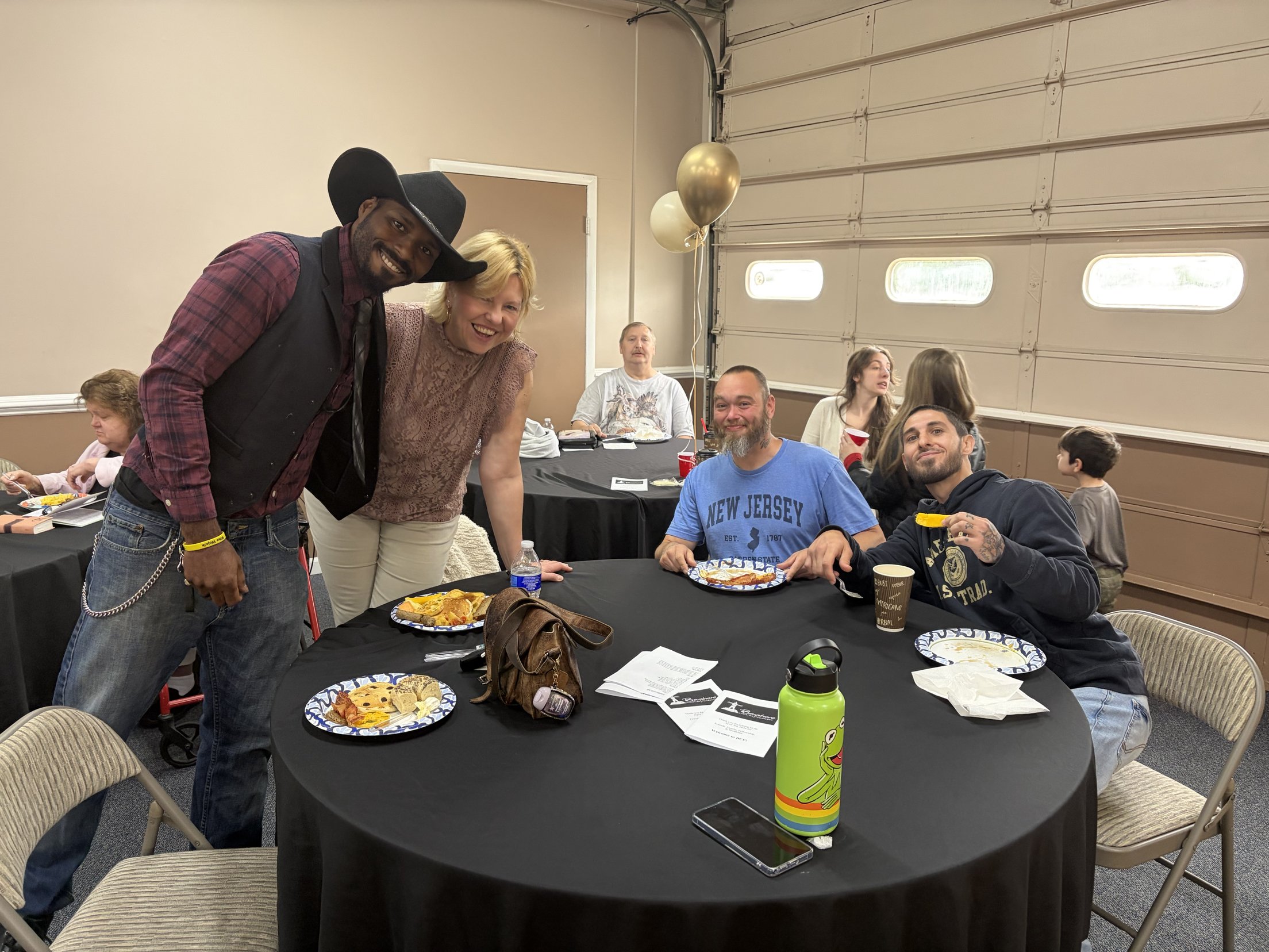 People gathered around a table at a party or celebration, with some posing and smiling for the camera. There are plates of food, drinks, and balloons in the background.