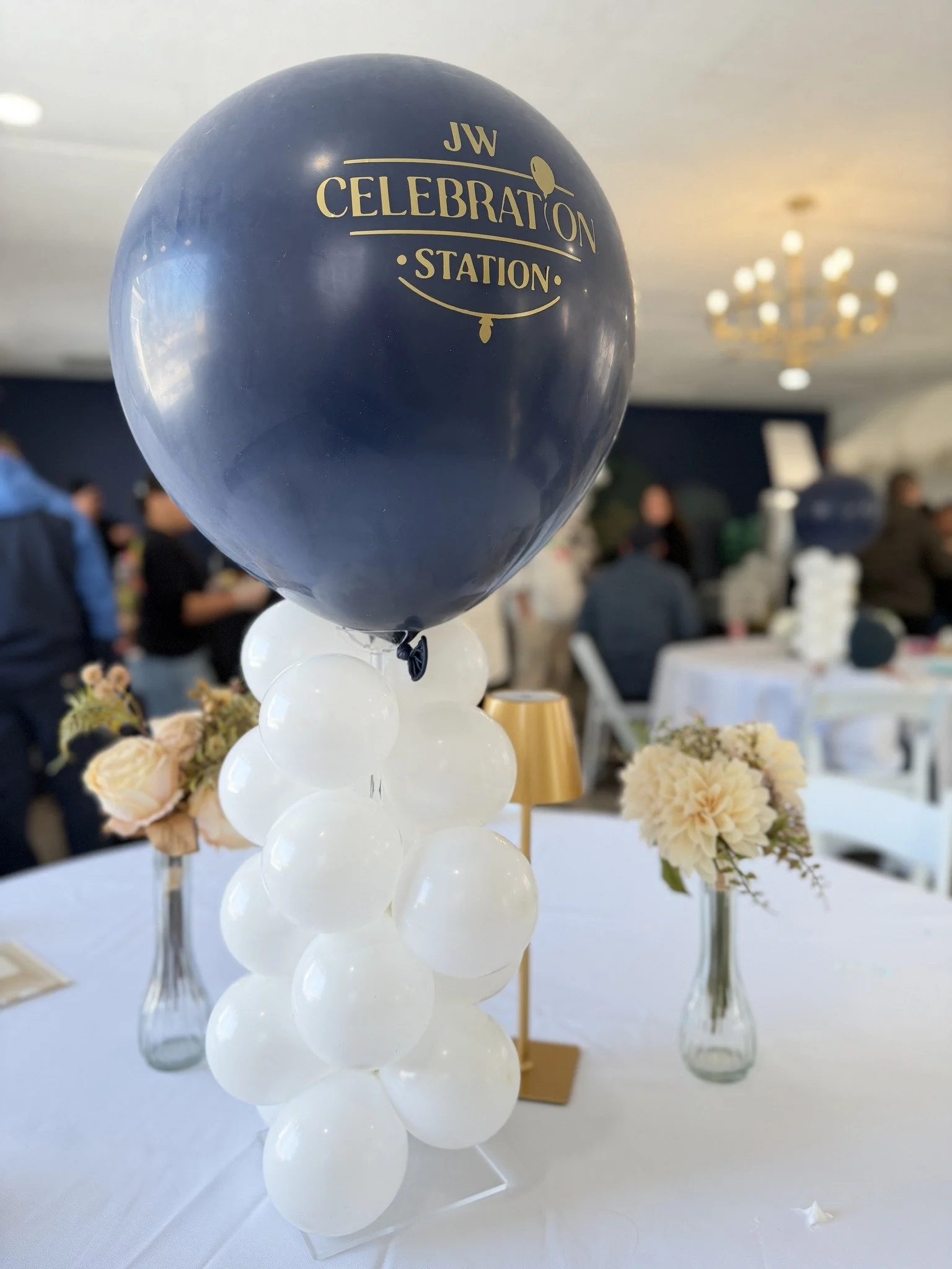Balloon centerpiece with navy blue and white balloons at a celebration event, with floral arrangements in vases and tables in background