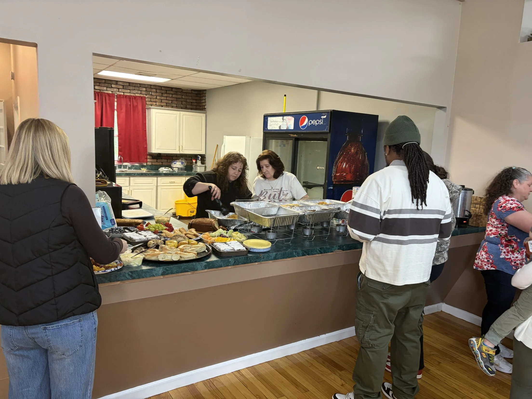 People serving and receiving food at a buffet-style setup in an indoor space with a kitchen in the background.