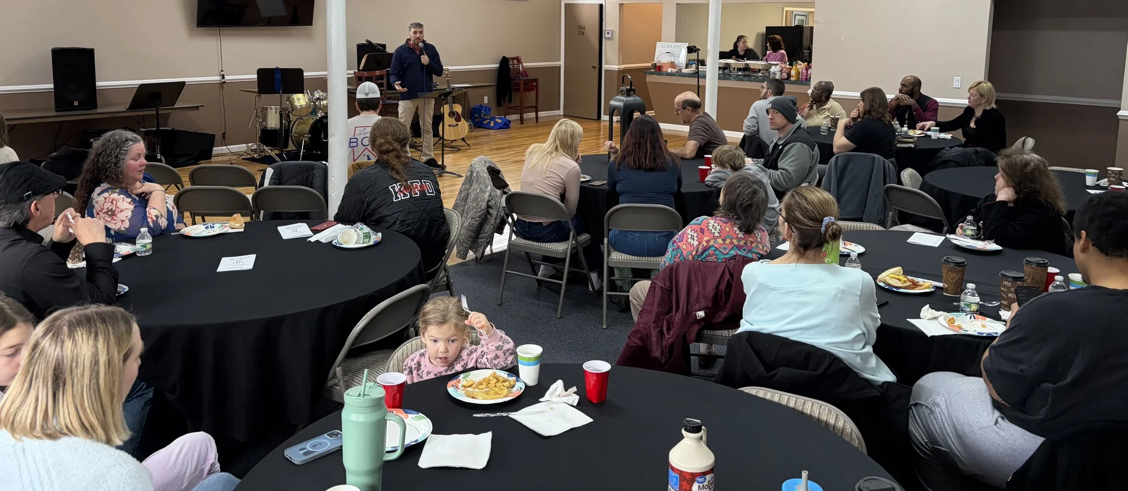 A group of people sitting at round tables in a banquet room, listening to a man speaking with a microphone. The tables have plates of food, cups, and bottles, and in the background, there is a small stage with musical instruments and a bar area.