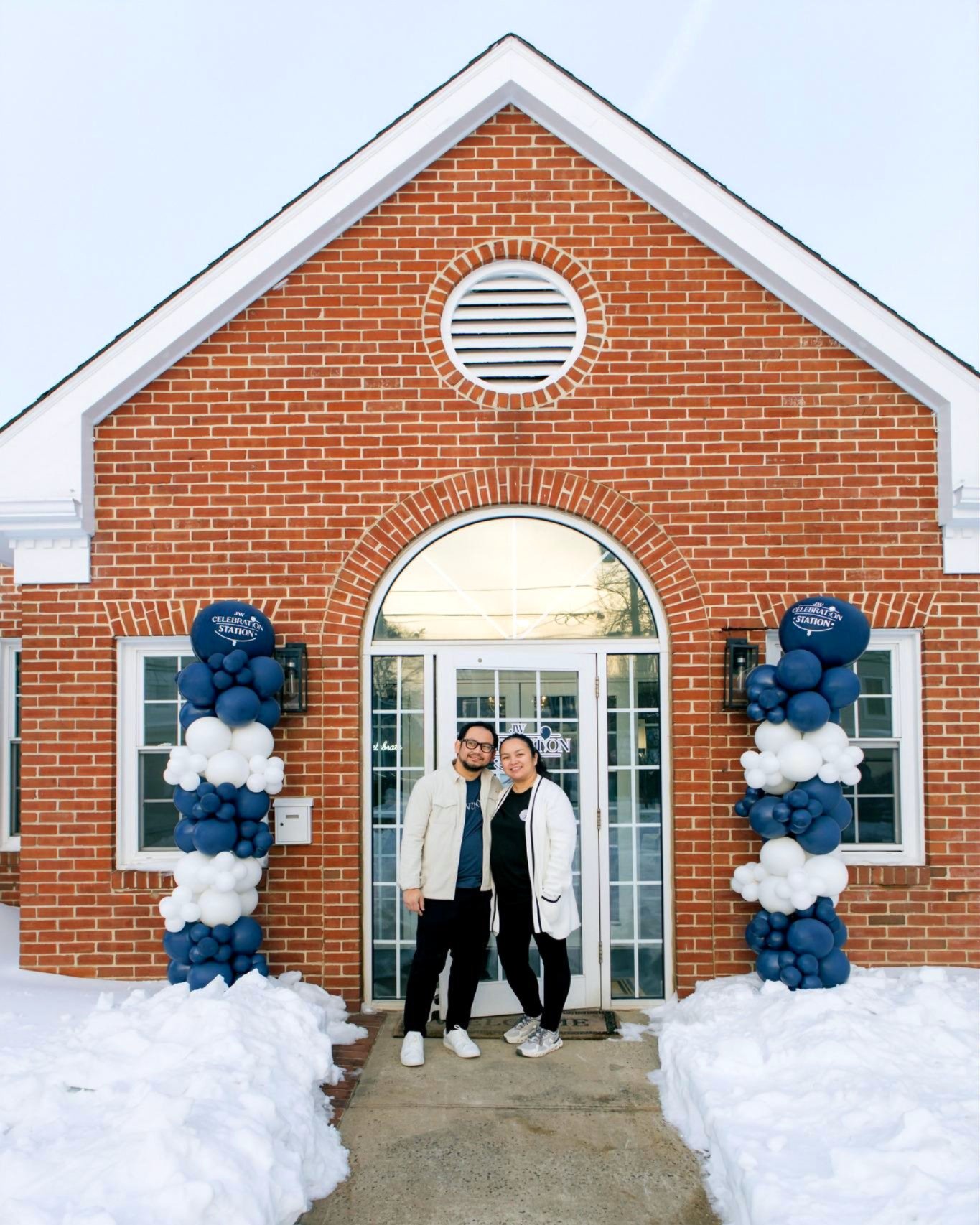 A couple standing in front of a brick building decorated with blue, white, and dark blue balloons for an event. Snow is on the ground.