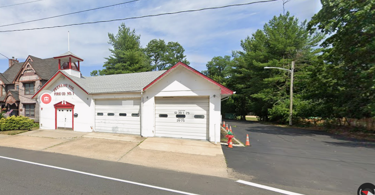 White fire station with two garage doors, red accents, a small tower on the roof, and a sign that says "FIRE CO NO. 1." There are orange traffic cones and a fire hydrant in front, with trees and houses in the background.
