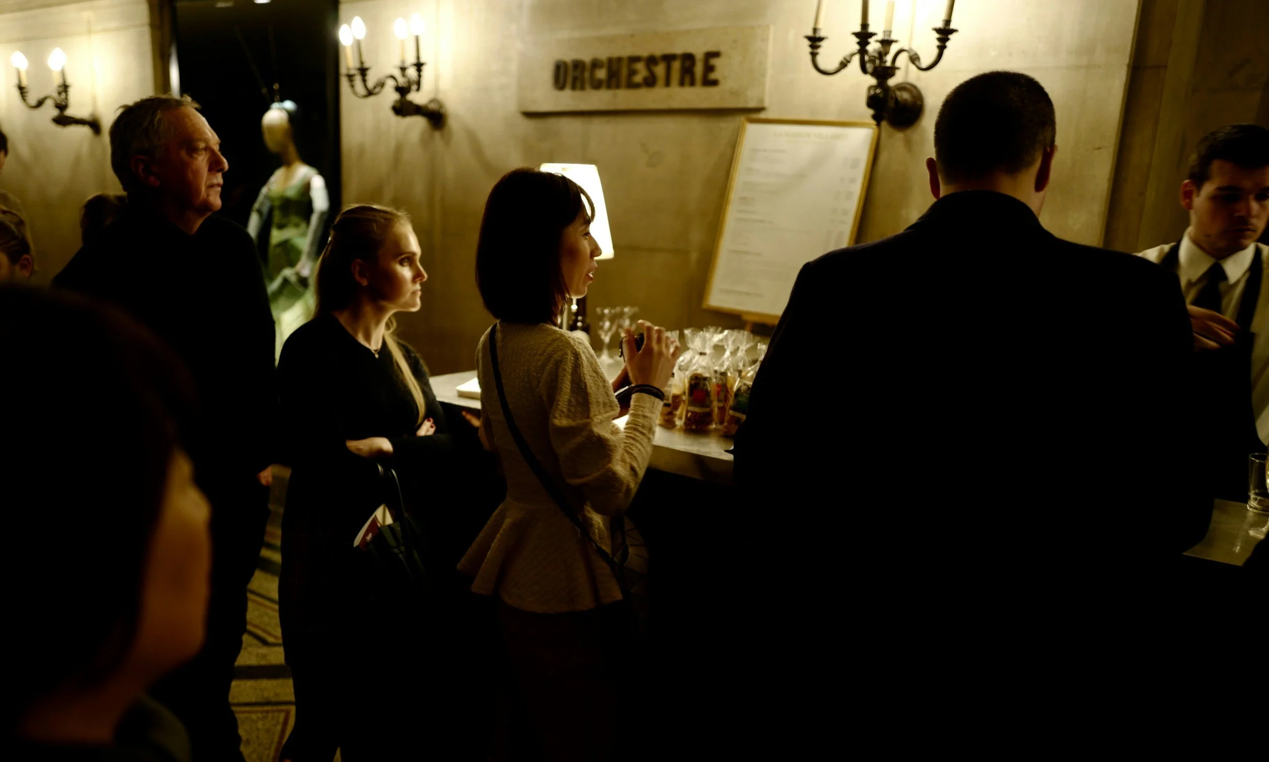 People standing at a reception desk in a room labeled 'ORCHESTRE', with warm lighting and decorative wall sconces.