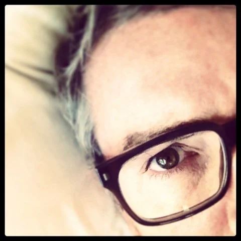 Close-up of a person's face with glasses and brown eyes, lying on a pillow.