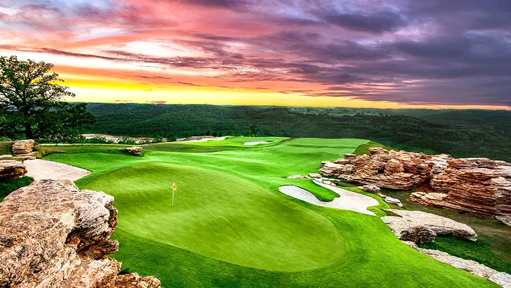 A scenic golf course at sunset with lush green fairways, sand traps, rocky formations, and a colorful sky.