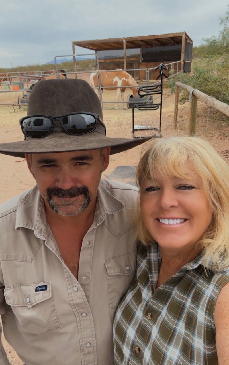A man wearing a large brown hat with sunglasses on top and a woman, with a horse in the background in a fenced outdoor area.