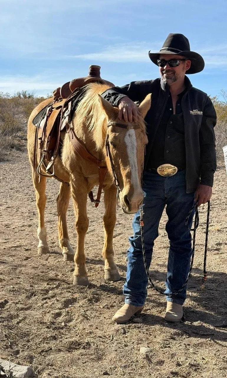 A man in cowboy attire, wearing a large black hat, sunglasses, a black jacket, and jeans, stands next to a light brown horse with a saddle, in a desert landscape with sparse vegetation and a blue sky.