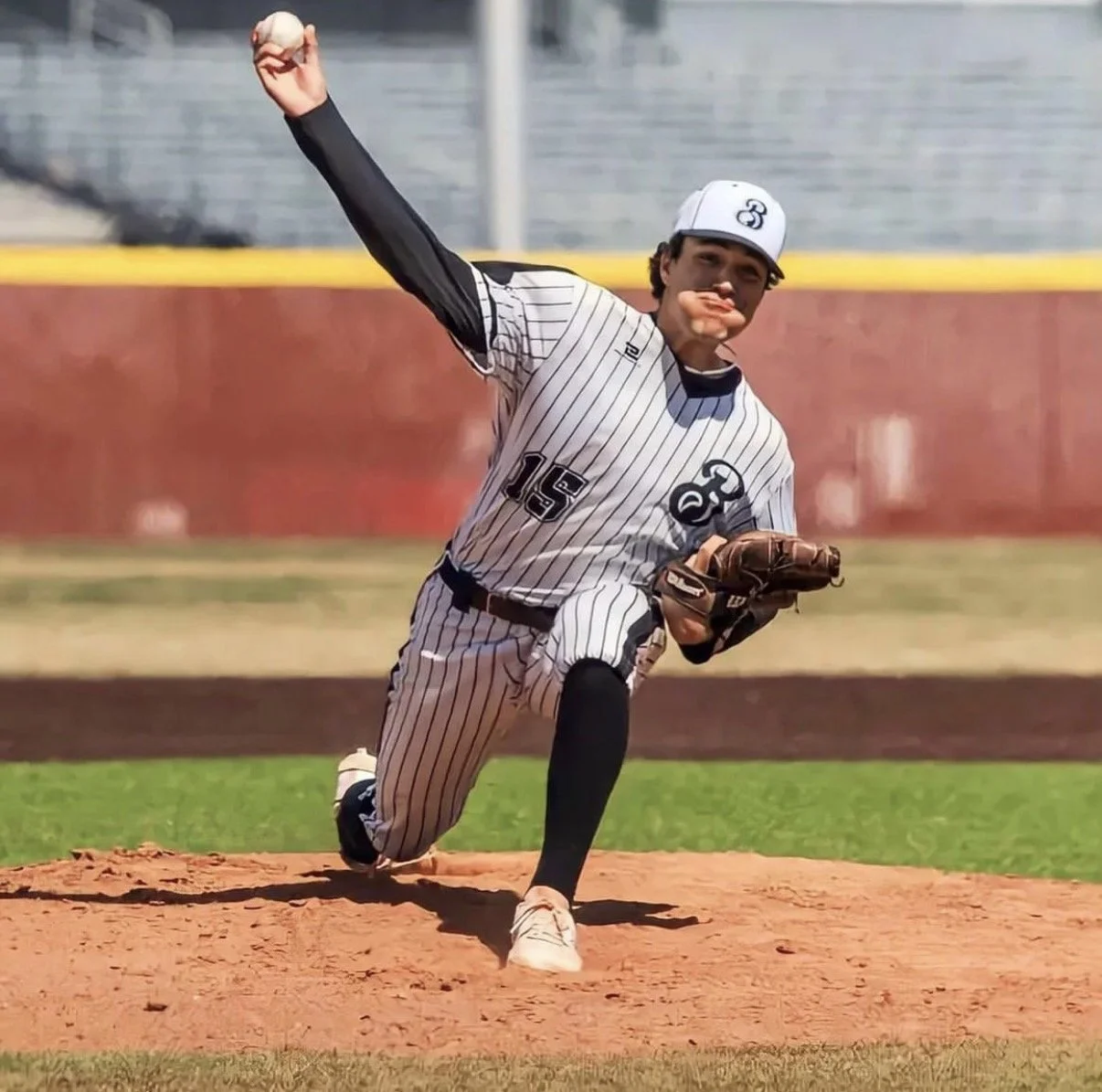 A young baseball player in a pinstripe uniform and cap is pitching on the mound, mid-throw, with his arm extended and glove on his left hand.