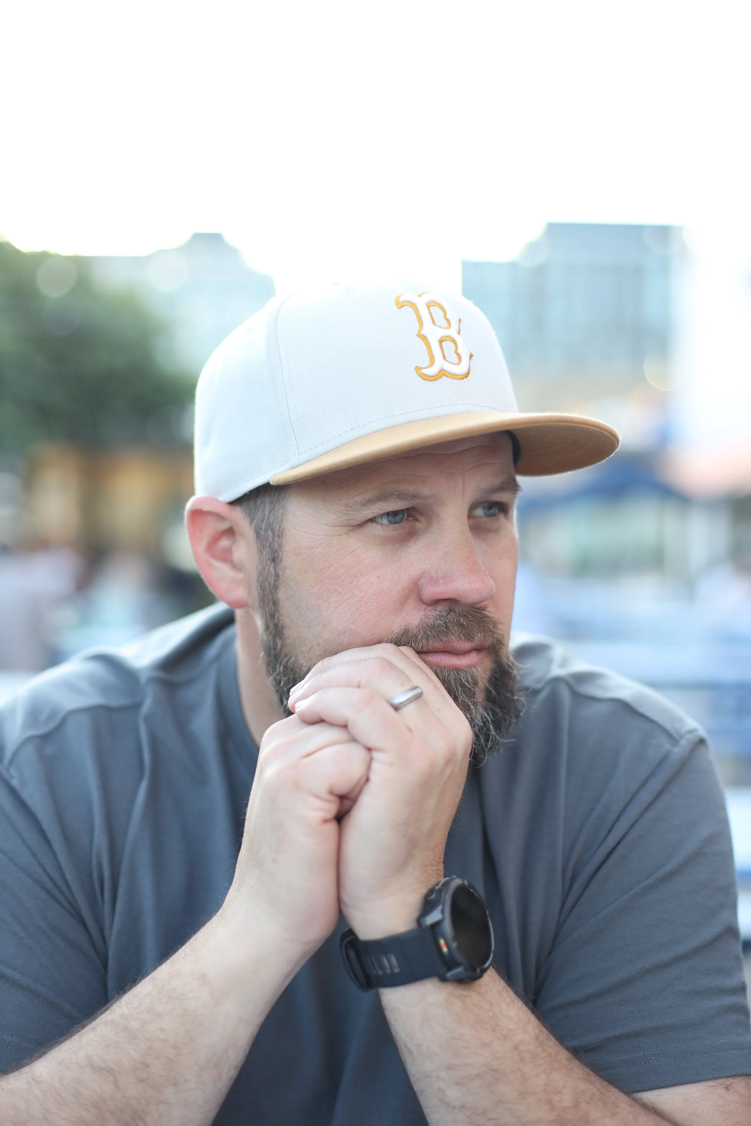 Close-up of a man with a beard wearing a white baseball cap with a yellow logo, resting his chin on his hands, outdoors in a city setting.