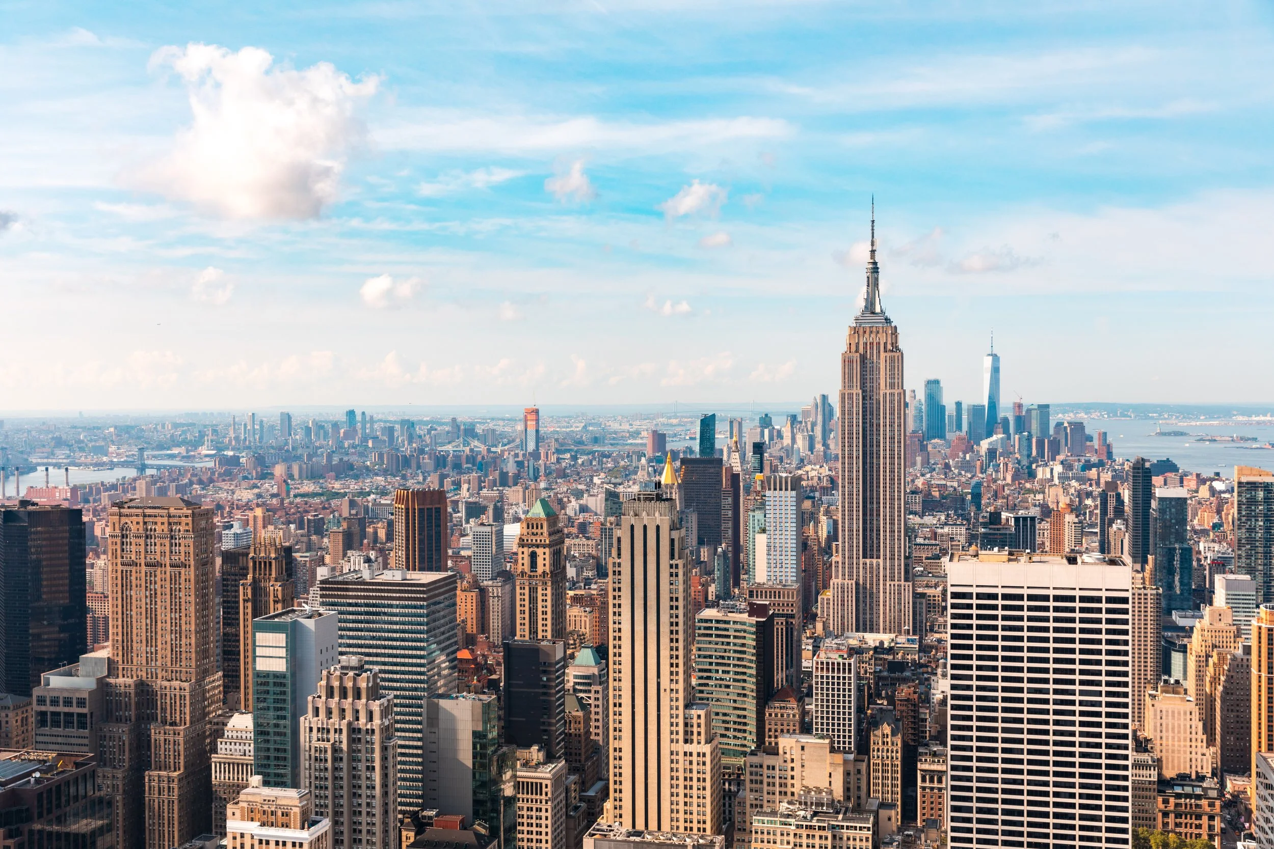 A panoramic view of Manhattan skyline featuring the Empire State Building in the foreground and One World Trade Center in the background, under a partly cloudy sky.