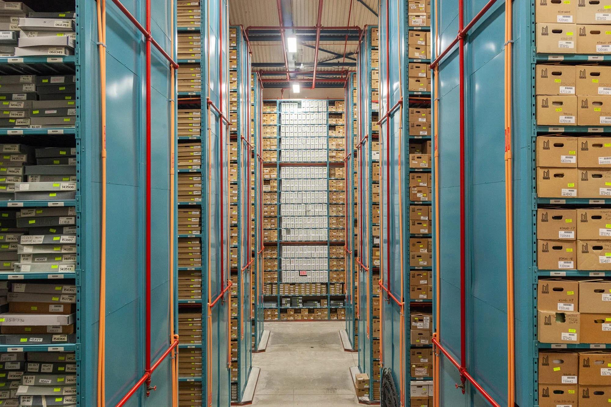 View of tall storage shelves filled with labeled boxes in a warehouse, with a narrow aisle and a cart on the floor.