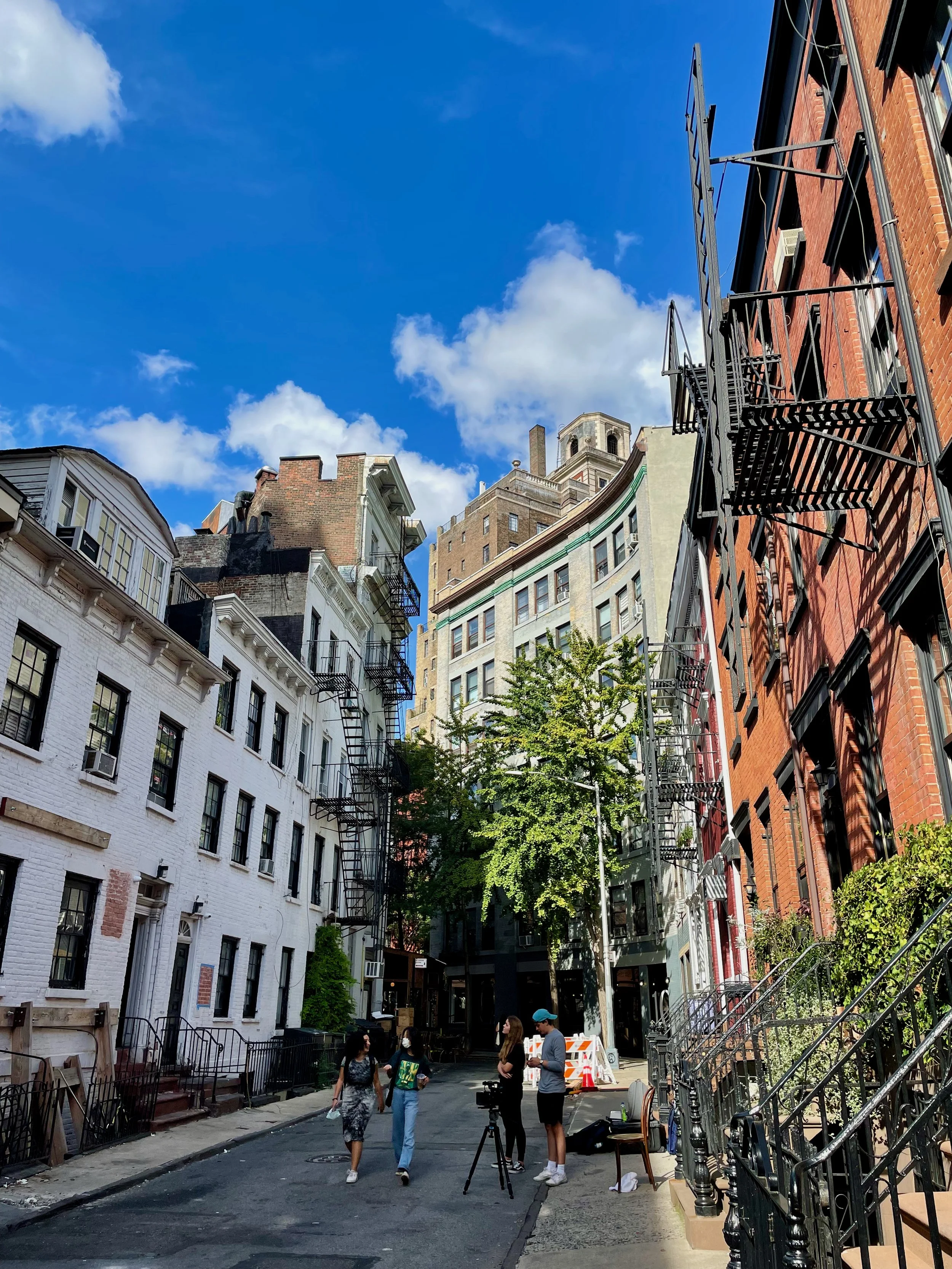 People standing on a city street surrounded by multi-story brick and white buildings under a blue sky with clouds, some bridge scaffolding, and trees.
