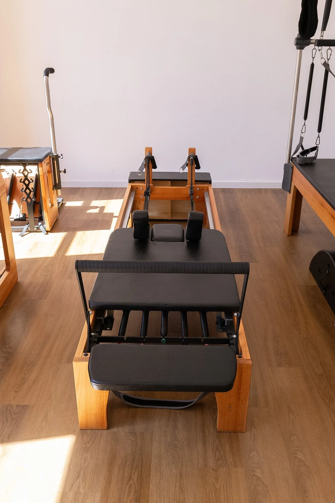 Uma sala de exercícios com equipamentos de pilates e academia de pilates, incluindo reformer, cadeiras de Pilates e acessórios em uma sala com piso de madeira e parede branca.