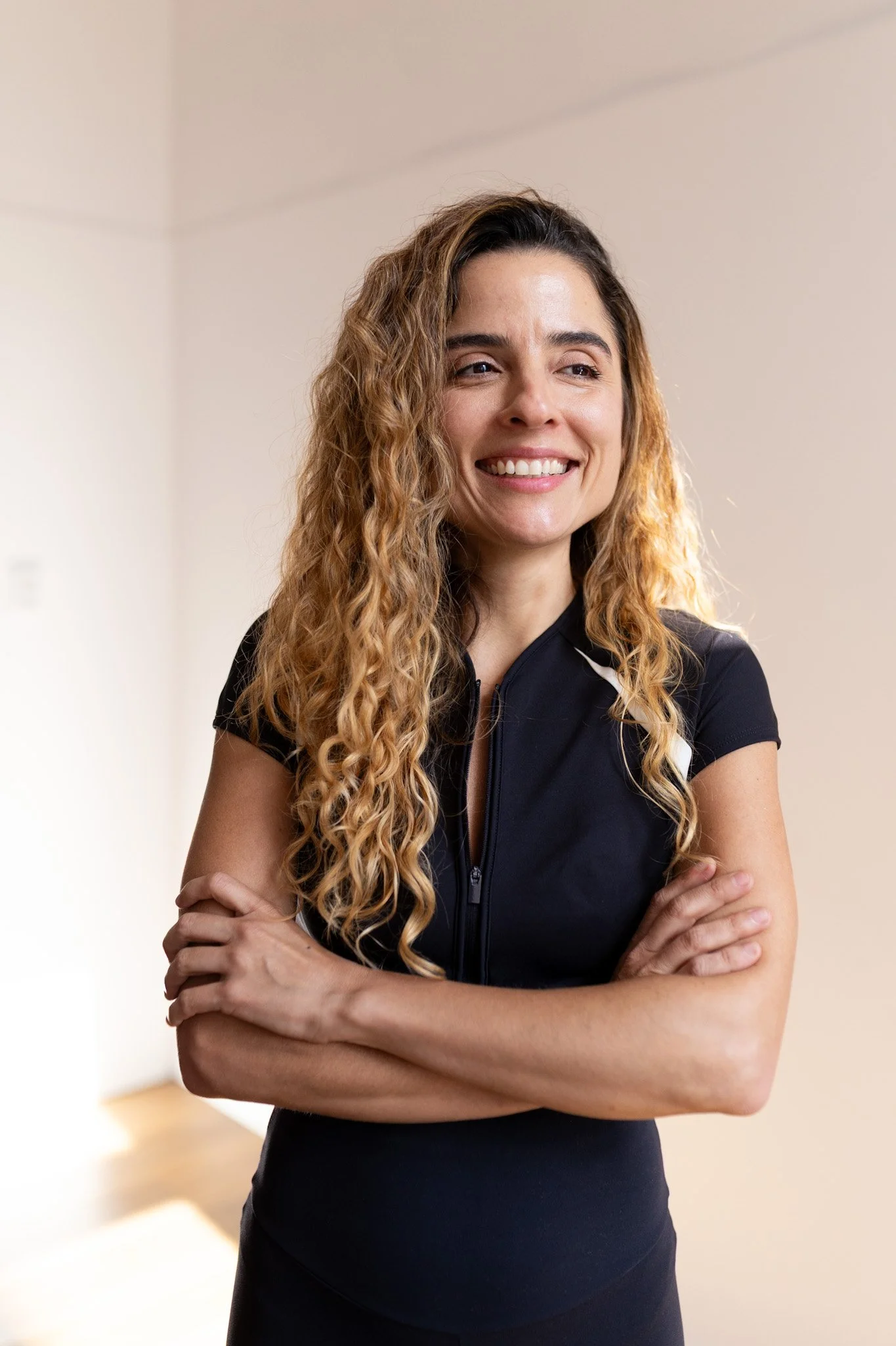 Mulher sorridente com cabelos cacheados e vestido preto, com os braços cruzados, em ambiente interno com parede branca ao fundo.