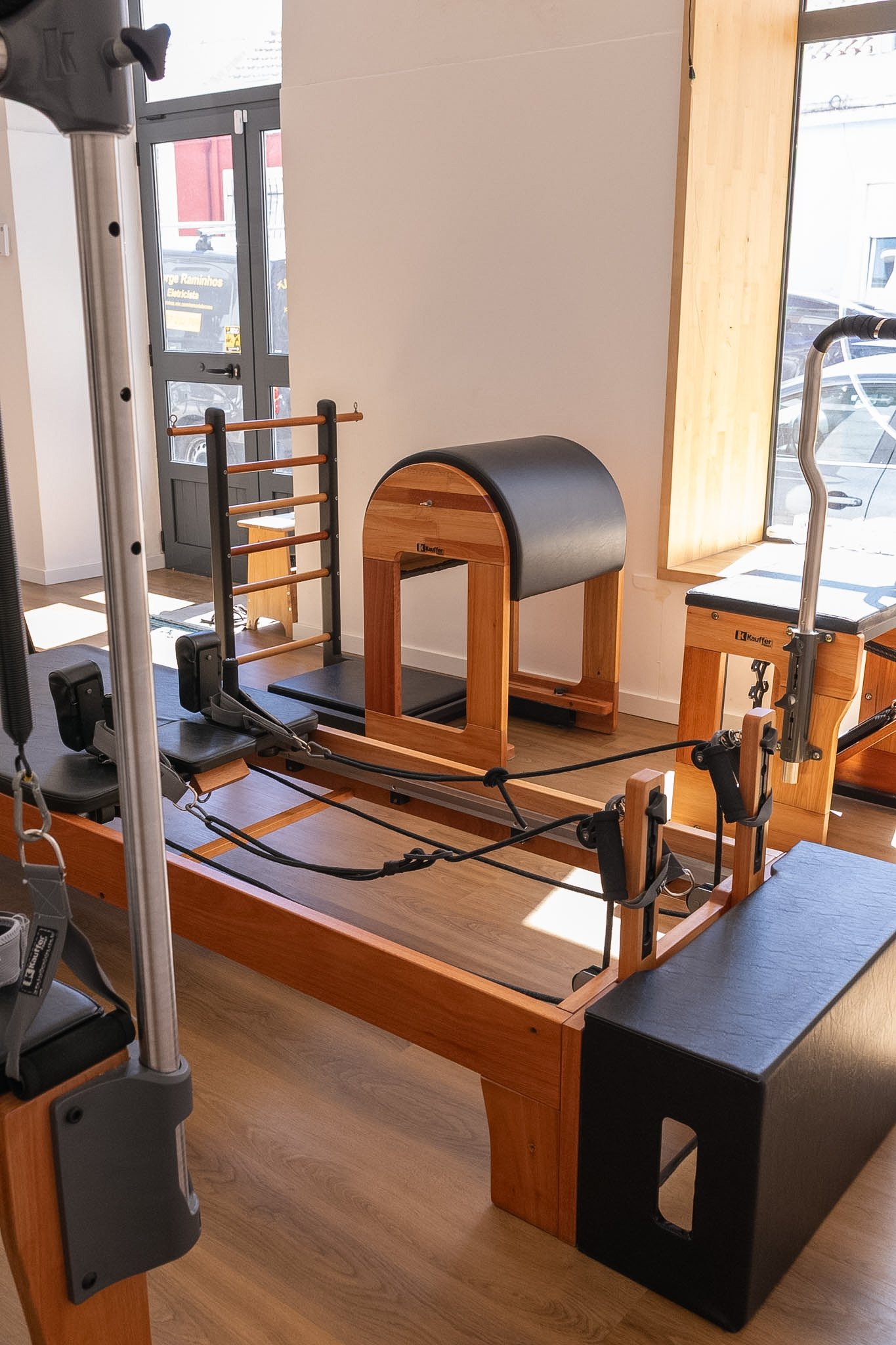 Ambiente de academia com equipamentos para exercícios de pilates, incluindo uma torre de resistência, uma mesa de cavalinho e plataformas de exercícios, com piso de madeira e uma grande janela de vidro.